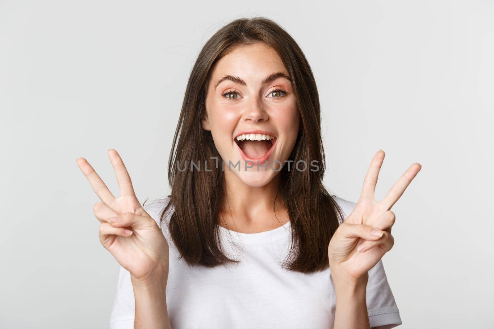 Portrait of beautiful and happy brunette girl smiling and showing peace gestures, standing white background excited.