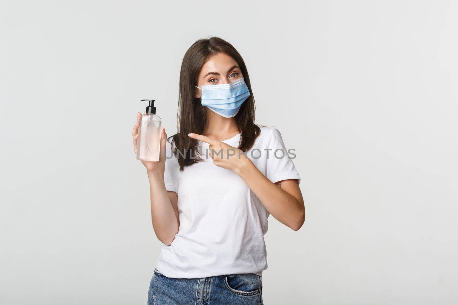 Covid-19, health and social distancing concept. Smiling cute brunette woman in medical mask advice using hand sanitizer, pointing at bottle.