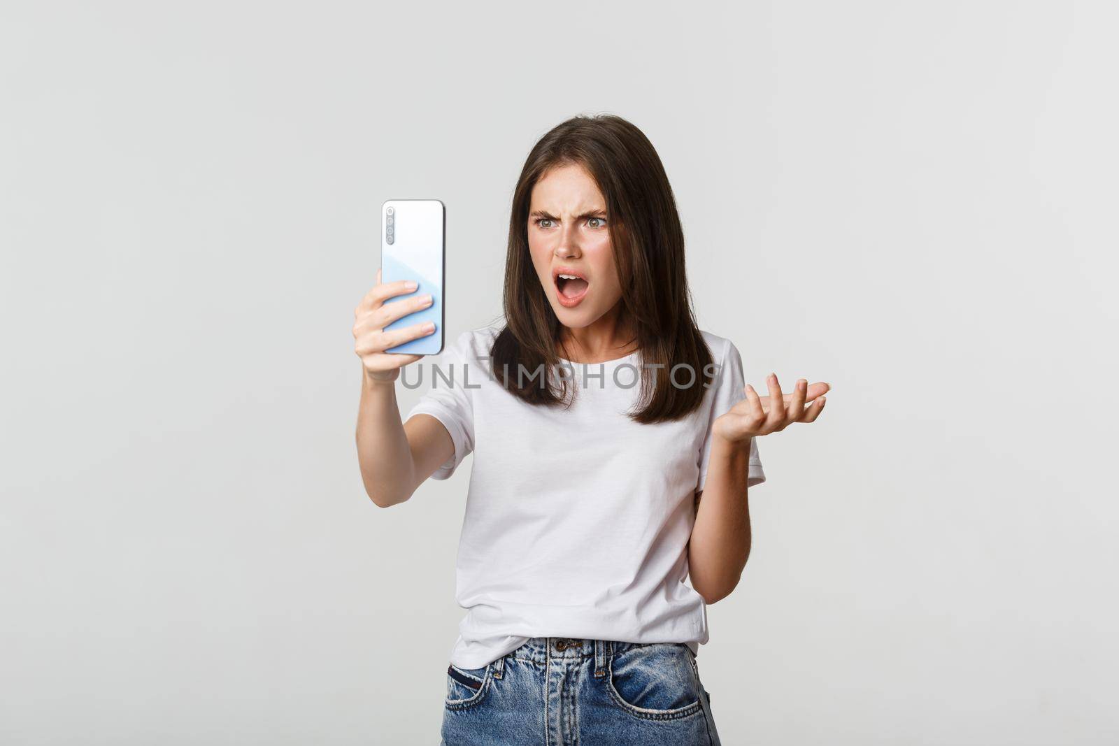 Frustrated young woman having argument on video call, holding smartphone, standing white background.