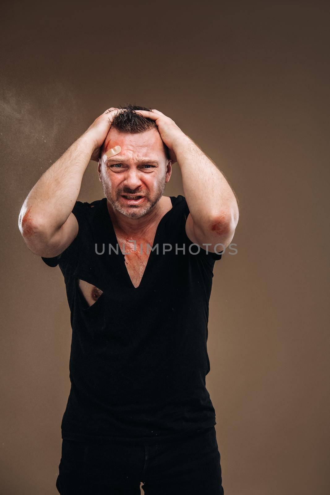 Against a gray background stands a battered angry man in a black T-shirt with wounds.