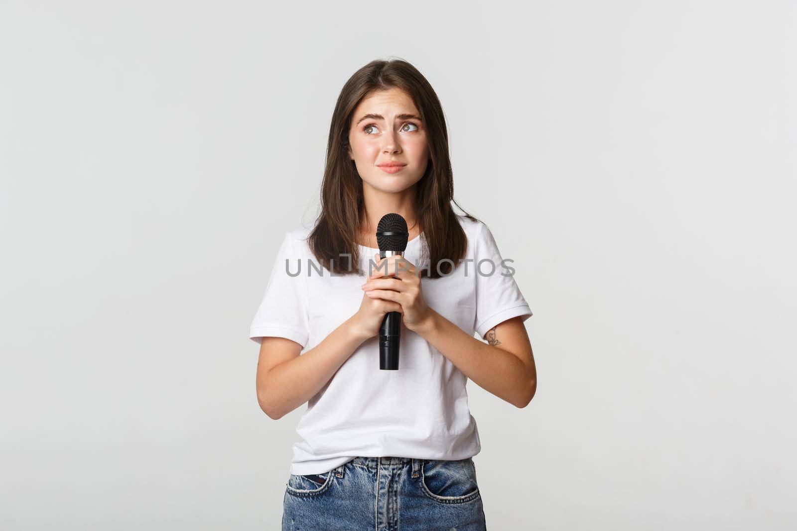 Shy cute brunette girl scared of singing in public, standing with microphone and looking nervous.
