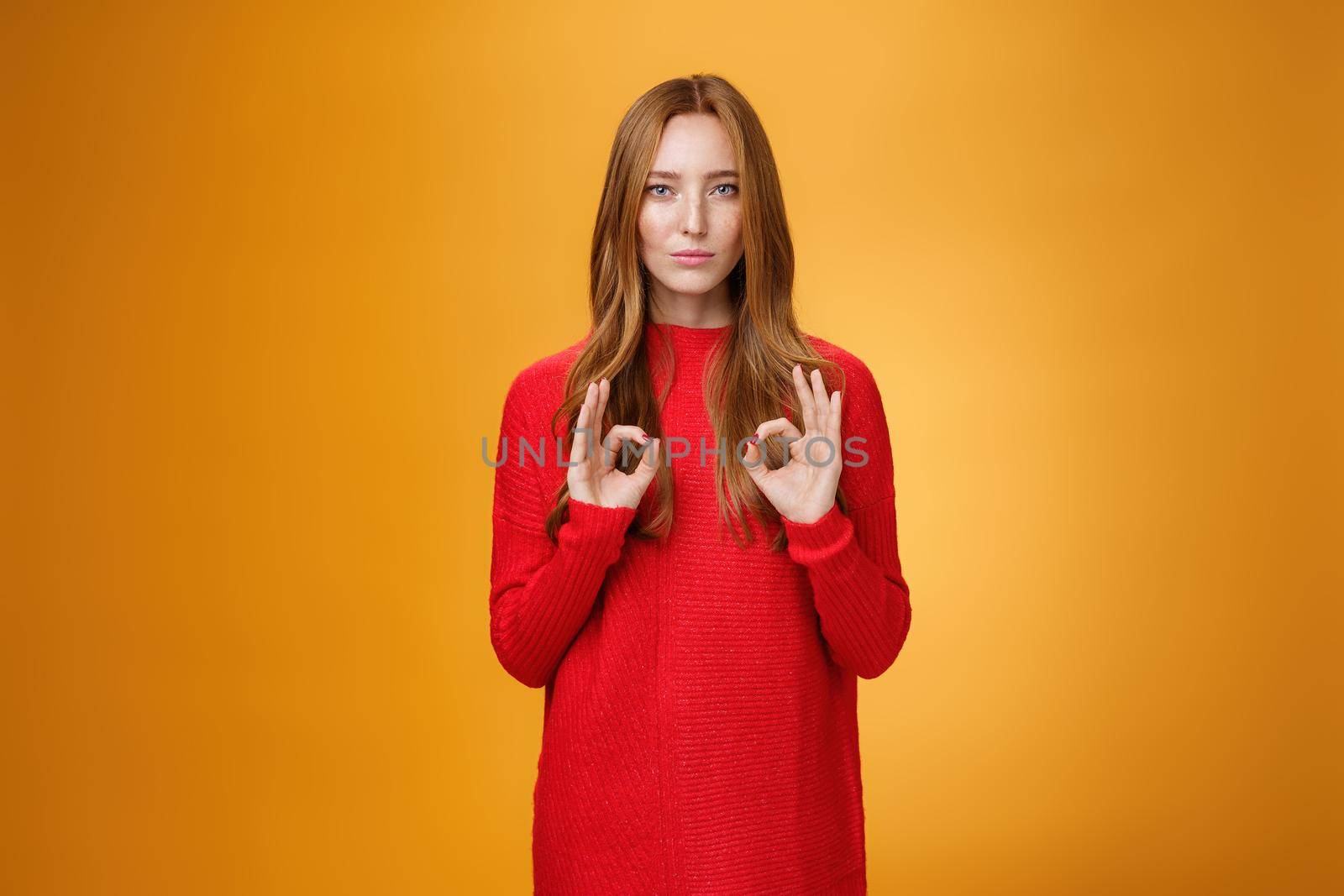 Calm and confident girl showing self-assurance and professionalism as making okay gesture with unemotional expression assuring everything goes by plan standing over orange background, giving approval.