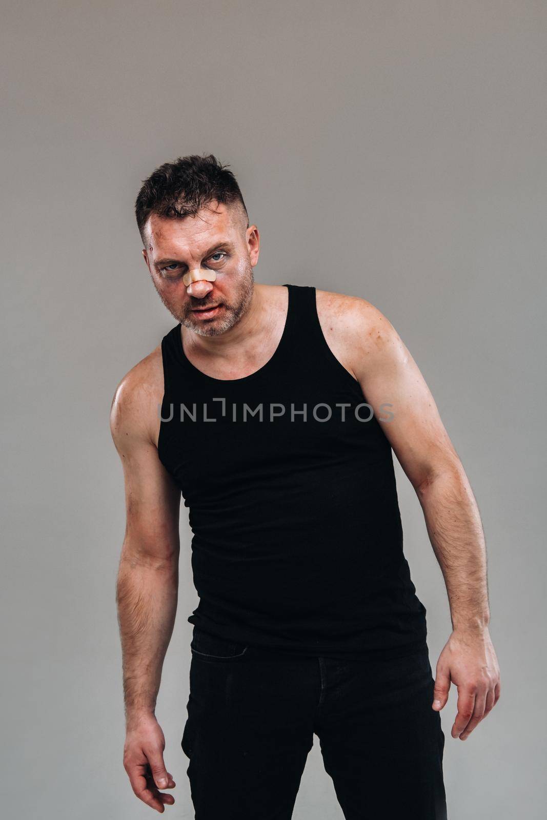 a battered man in a black T shirt who looks like a drug addict and a drunk stands against a gray background.