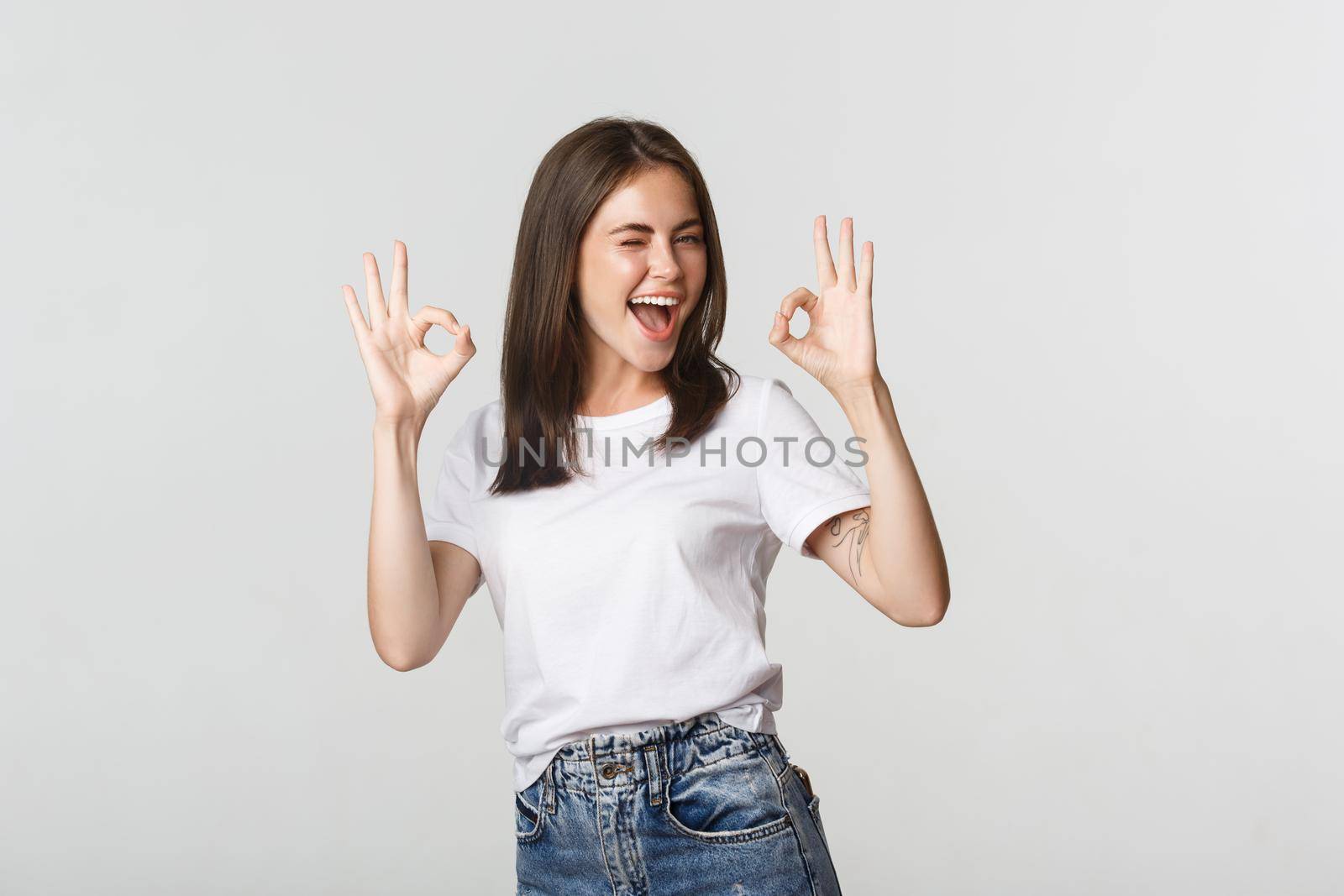 Satisfied cheerful brunette girl winking sassy and showing okay gesture in approval.