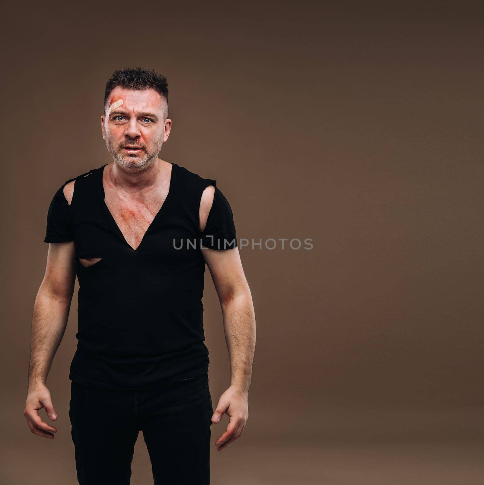 Against a gray background stands a battered angry man in a black T-shirt with wounds.