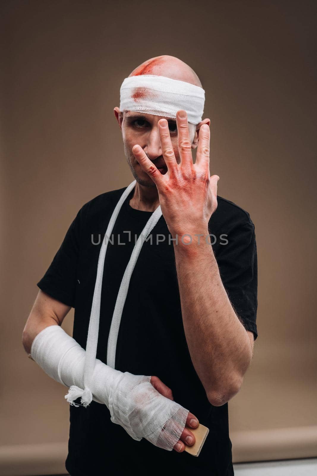 A battered man with a bandaged head and a cast on his arm stands on a gray background.