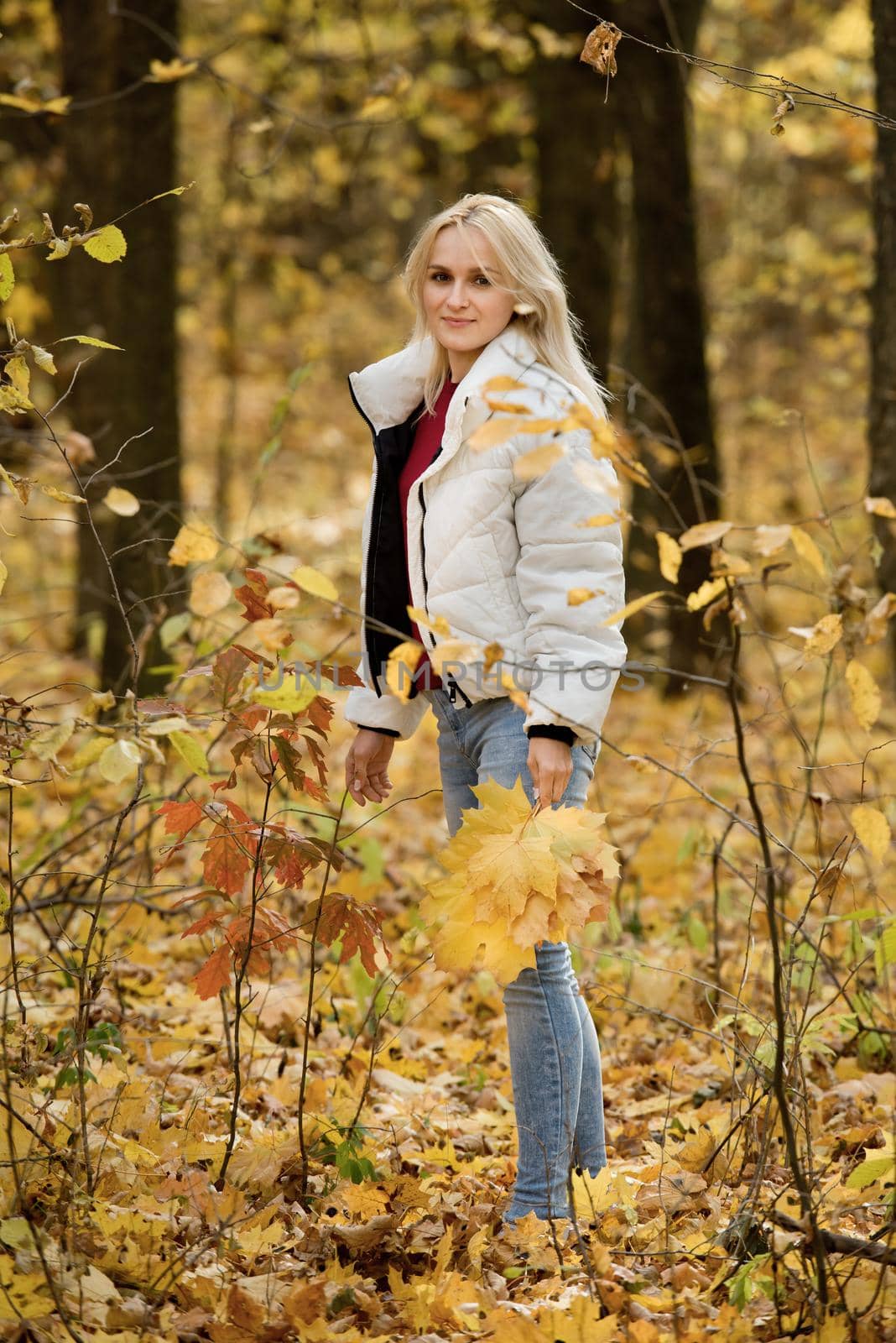Young blonde woman in the autumn forest, with a bouquet of yellow leaves in her hands.