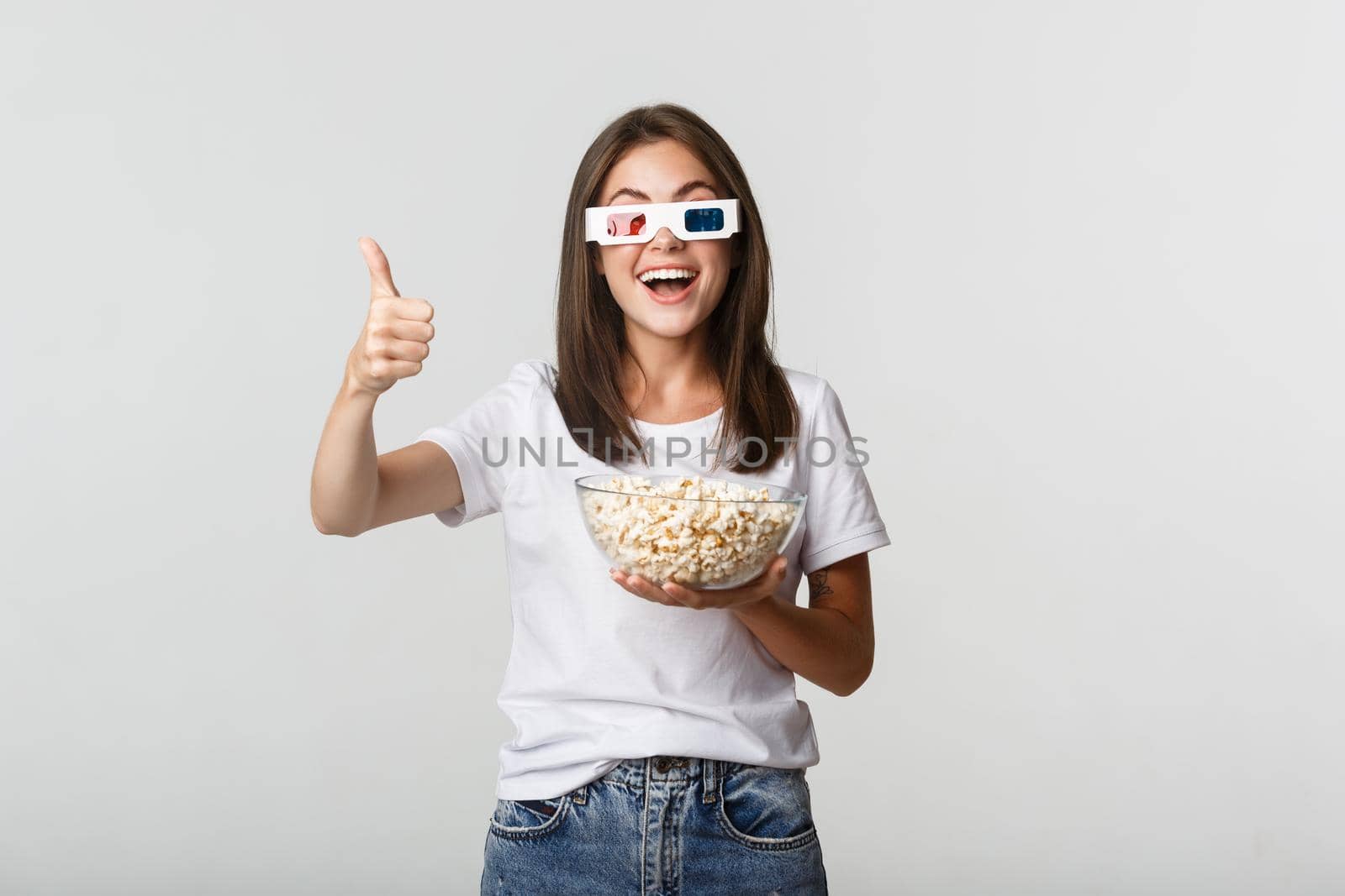 Portrait of satisfied happy girl in 3d glasses, holding bowl with popcorn and showing thumbs-up, like great tv series or movie.