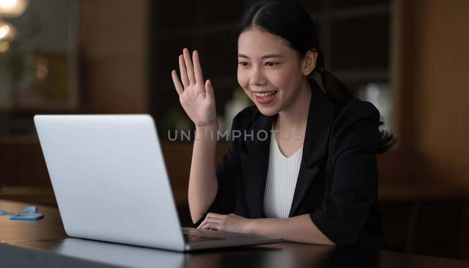 Image of happy asian woman wearing white shirt smiling and waving hand at laptop, while speaking or chatting on video call in office