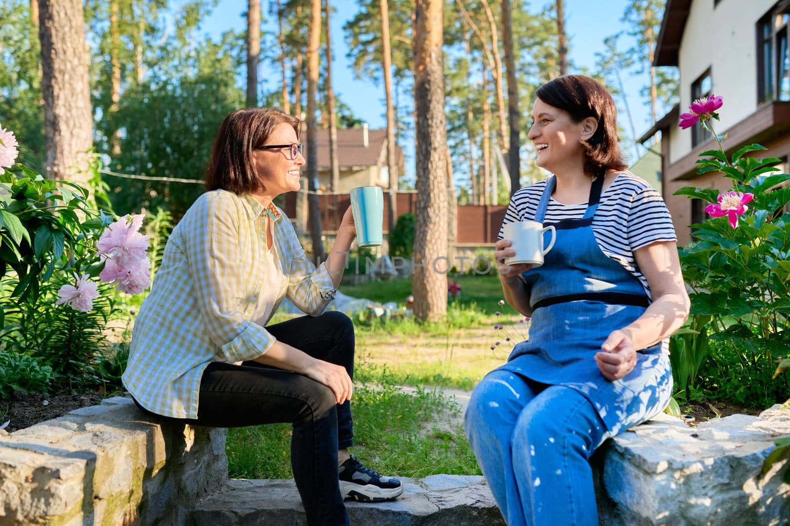 40s people, women, leisure, communication, friendship concept. Two laughing happy middle aged women sitting outdoors together in a spring garden, backyard with mugs of tea.