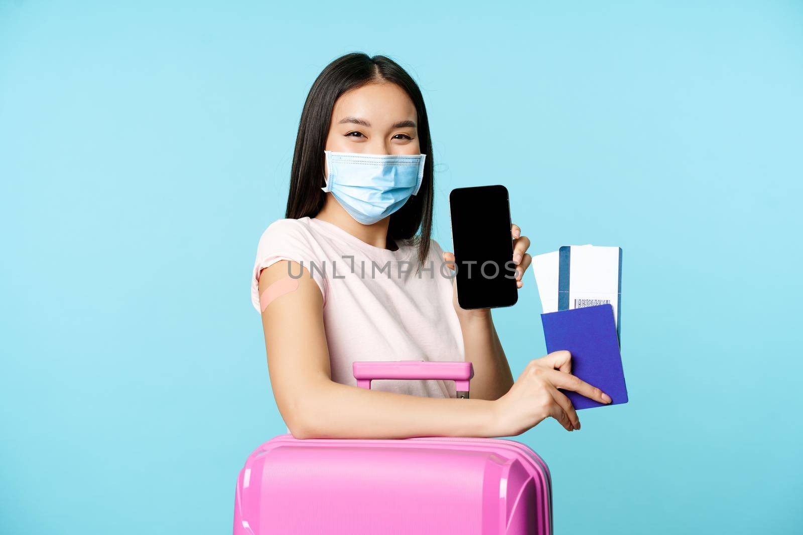 Tourism and healthcare concept. Asian female tourist with suitcare, wearing medical face mask for flight, showing smartphone screen and passport with tickets, vaccination certificate.