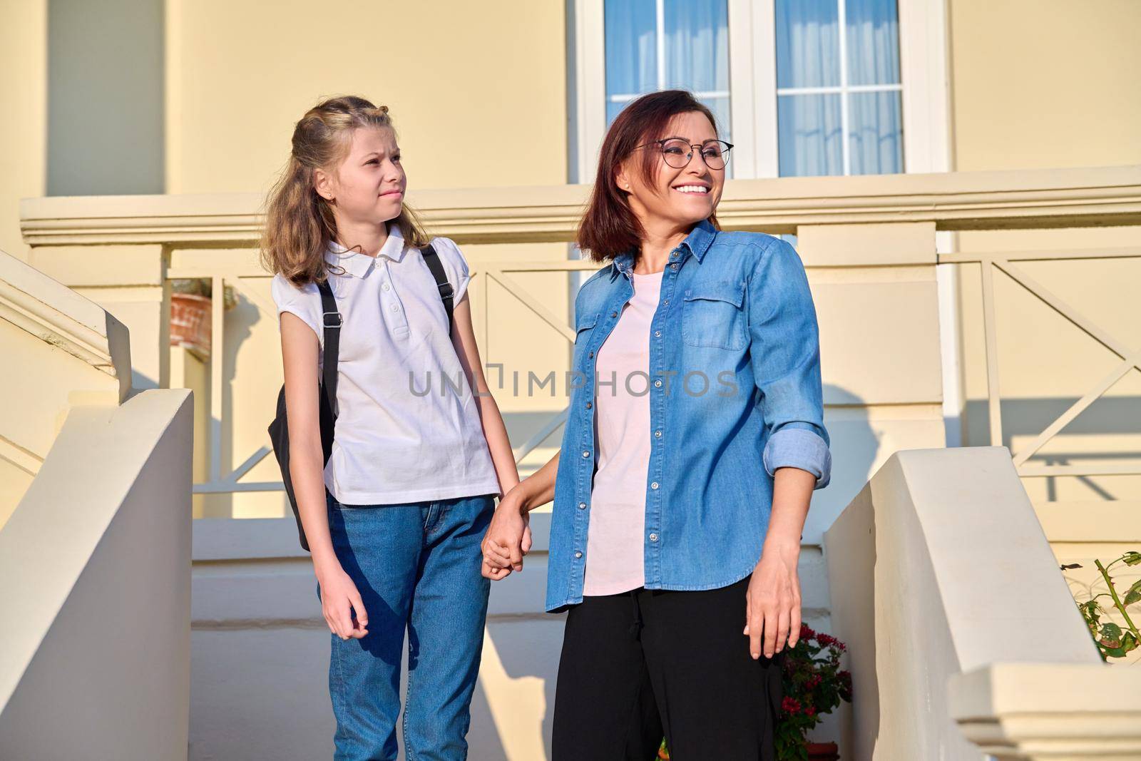 Mother and daughter schoolgirl with backpack on porch of house. Mom holding preteen student by hand as she goes to school on sunny morning. Parent child relationship, family, back to school concept