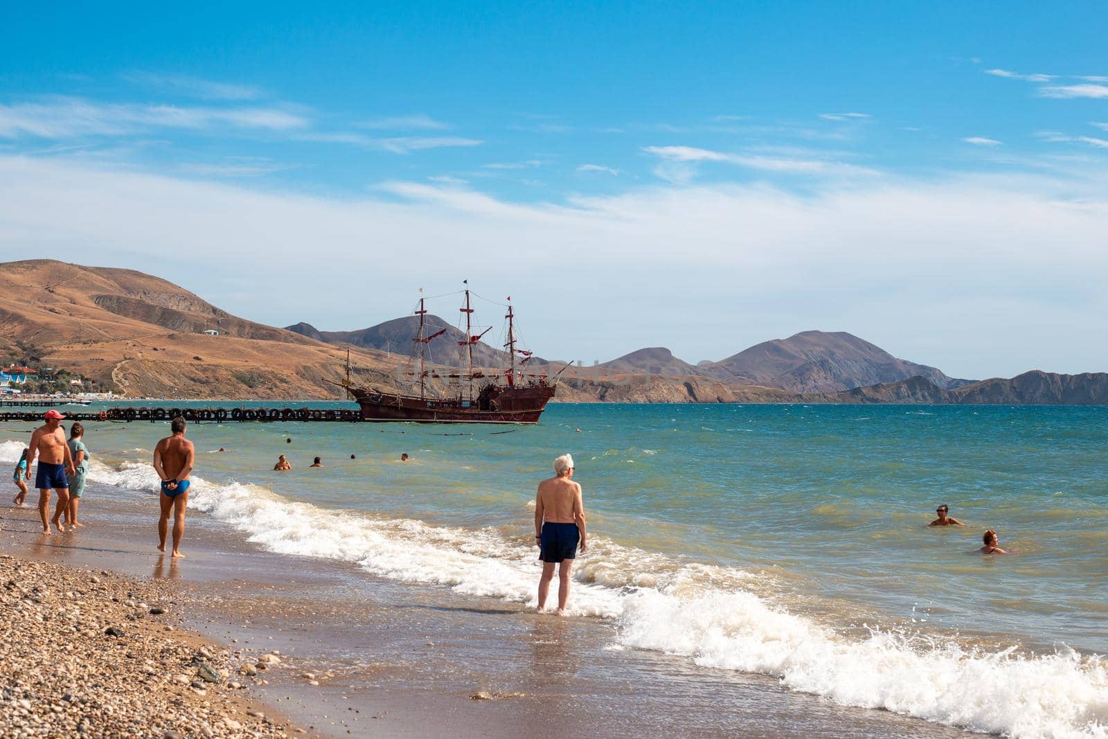tourists walk along a pebble beach on the Black Sea, where a ship is at the pier. Travel and tourism. Crimea, Koktebel-09.18.2021