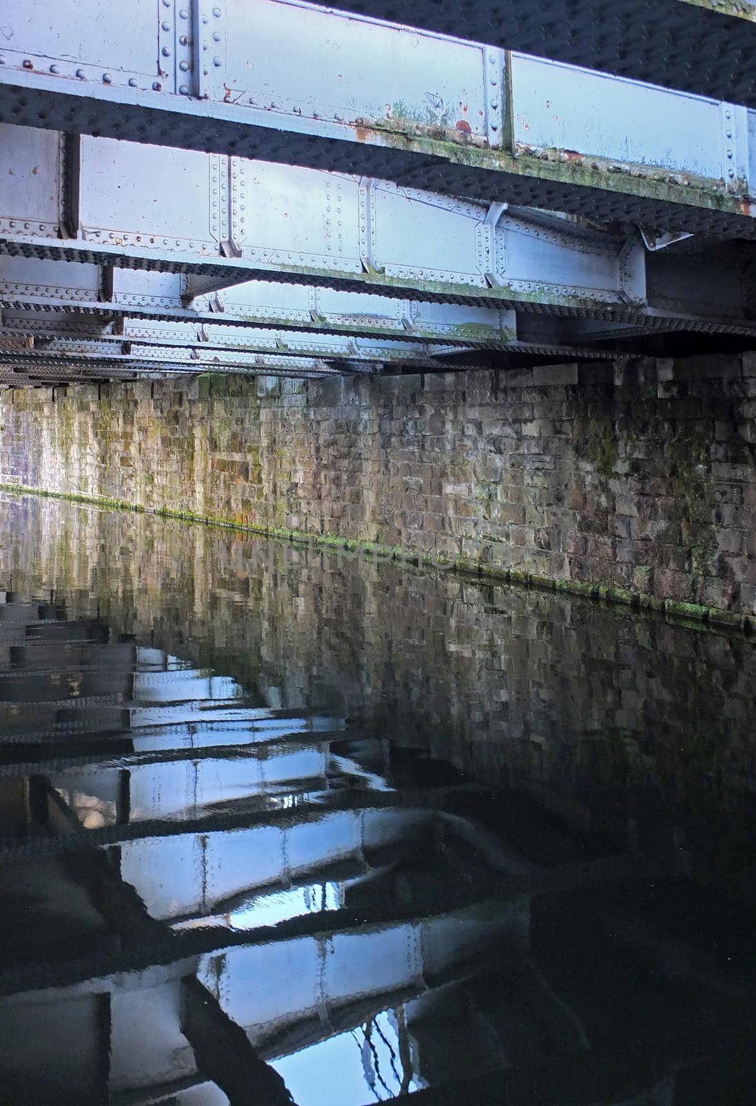 old steel girders reflected reflected in the water of a dark canal under a bridge