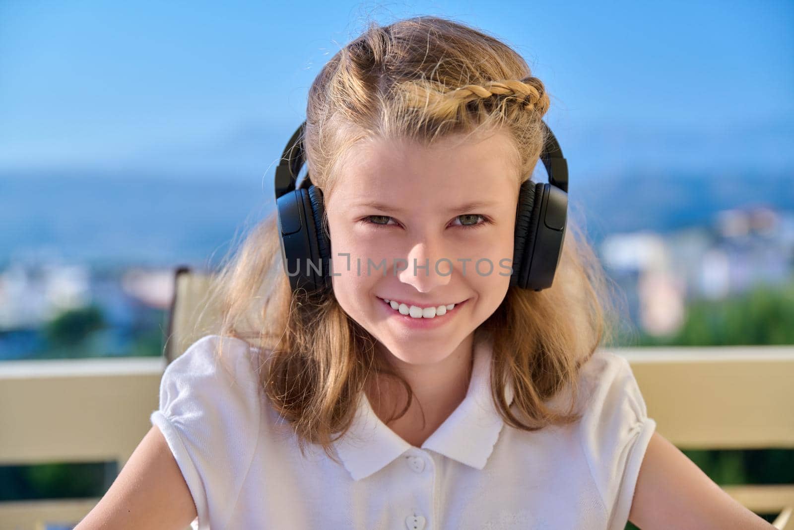 Head shot, portrait of child girl looking outdoors at camera. Preteen in headphones, smiling blonde, face close up, young female student, blue sunny sky background