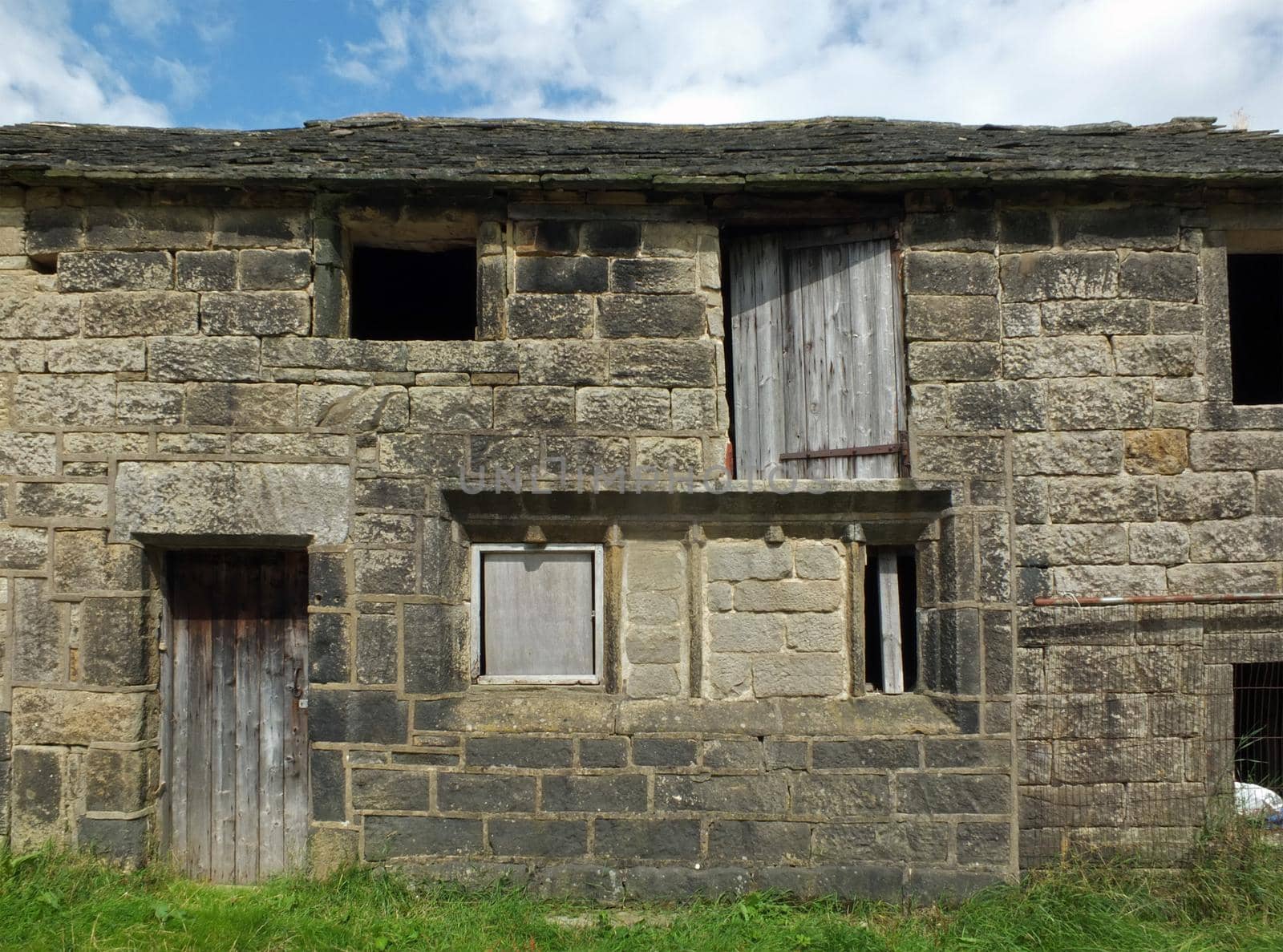 ancient abandoned stone cottage in a row of rural buildings with empty windows and wooden doors with the pavement overgrown with grass by philopenshaw