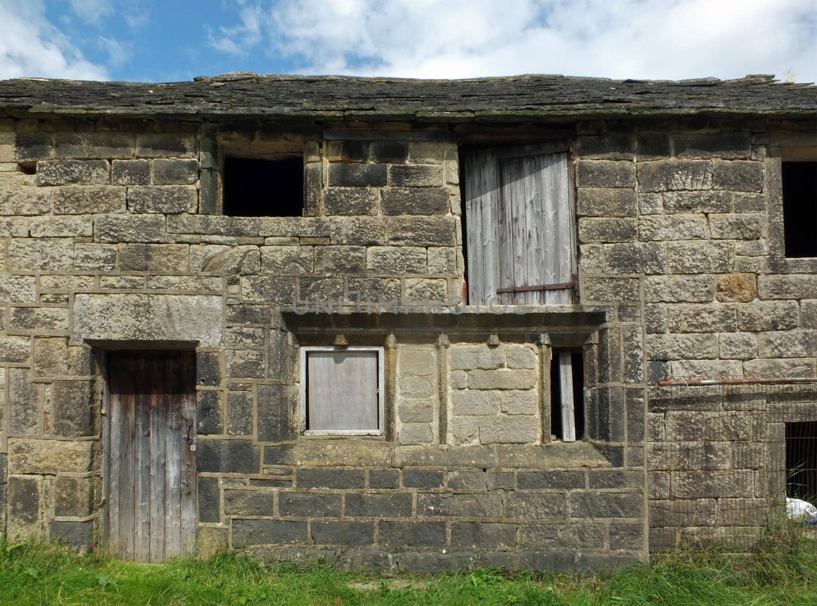 ancient abandoned stone cottage in a row of rural buildings with empty windows and wooden doors with the pavement overgrown with grass