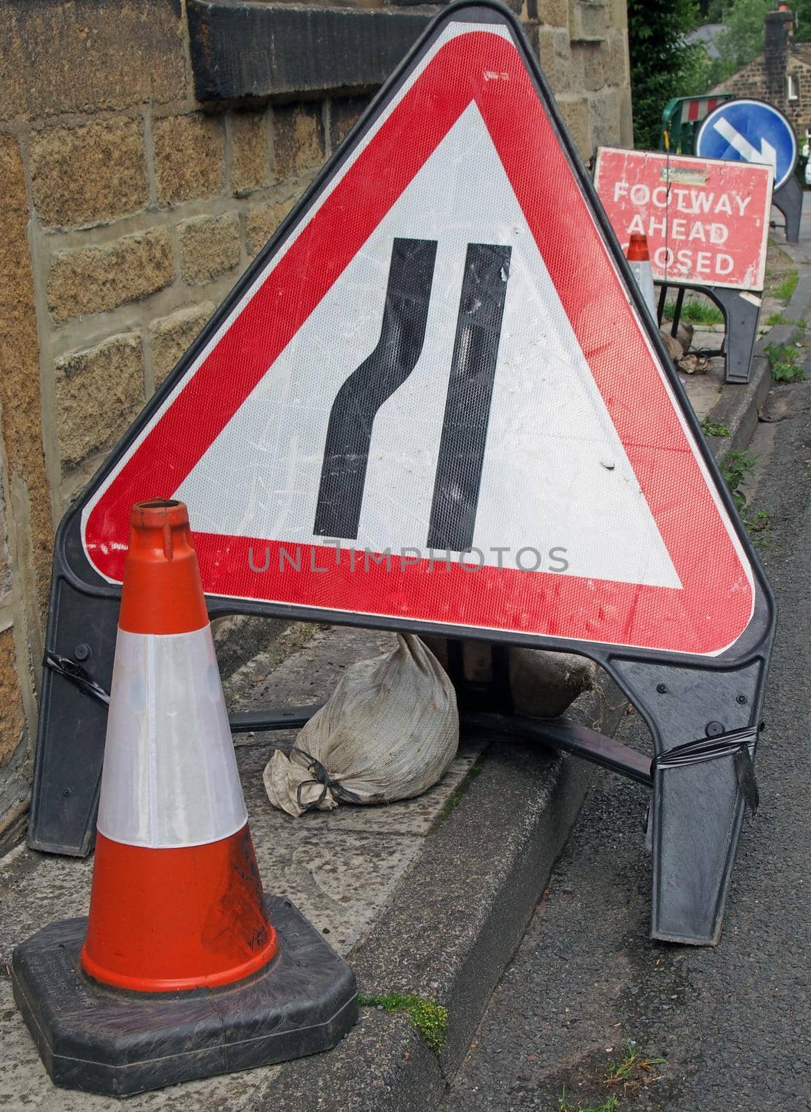a clutter of temporary warning and direction signs at the side of a narrow country road while repair work is being carried out