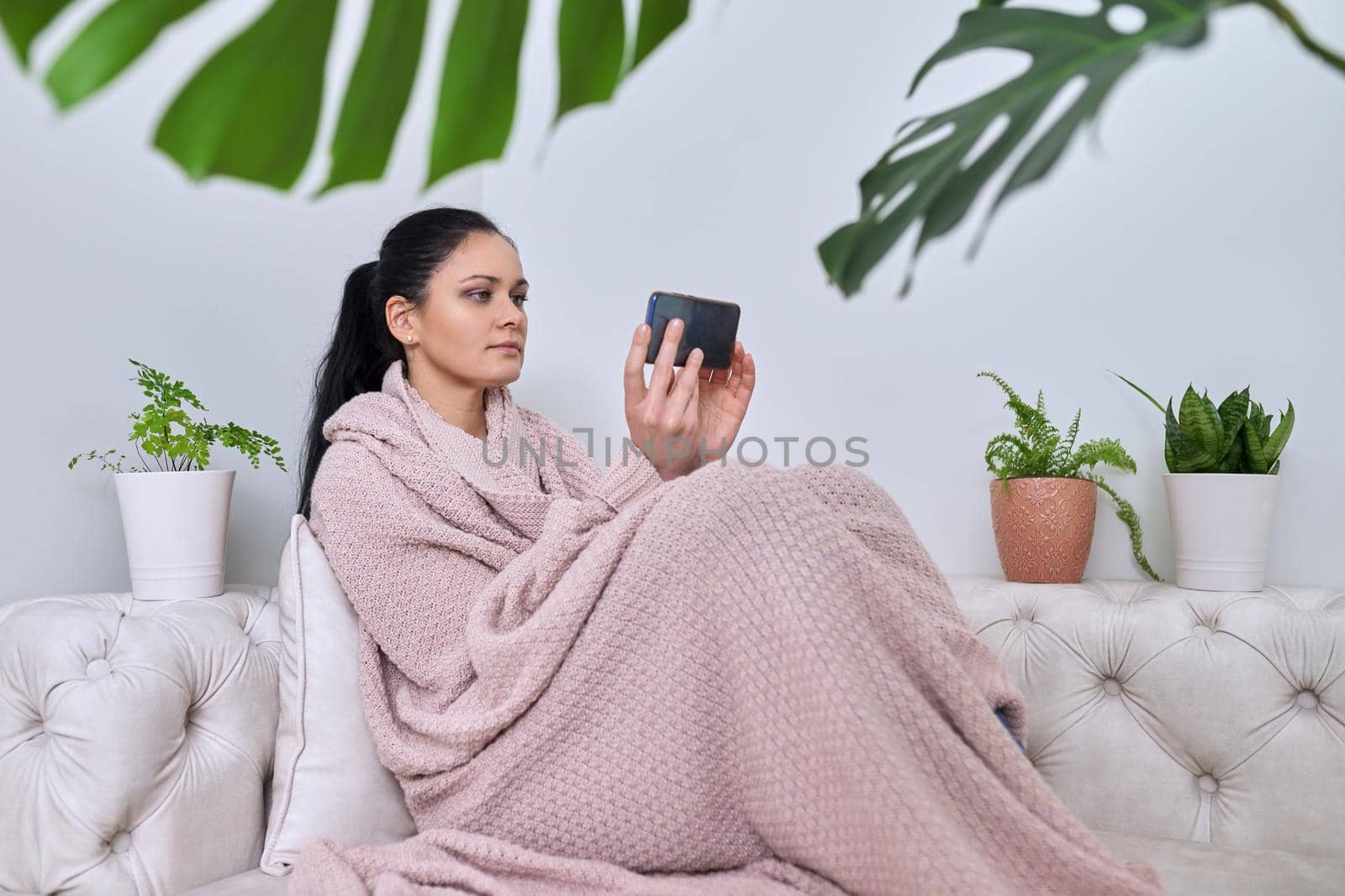 Middle-aged woman having a rest at home in winter fall season. A female under a warm knitted blanket with a smartphone