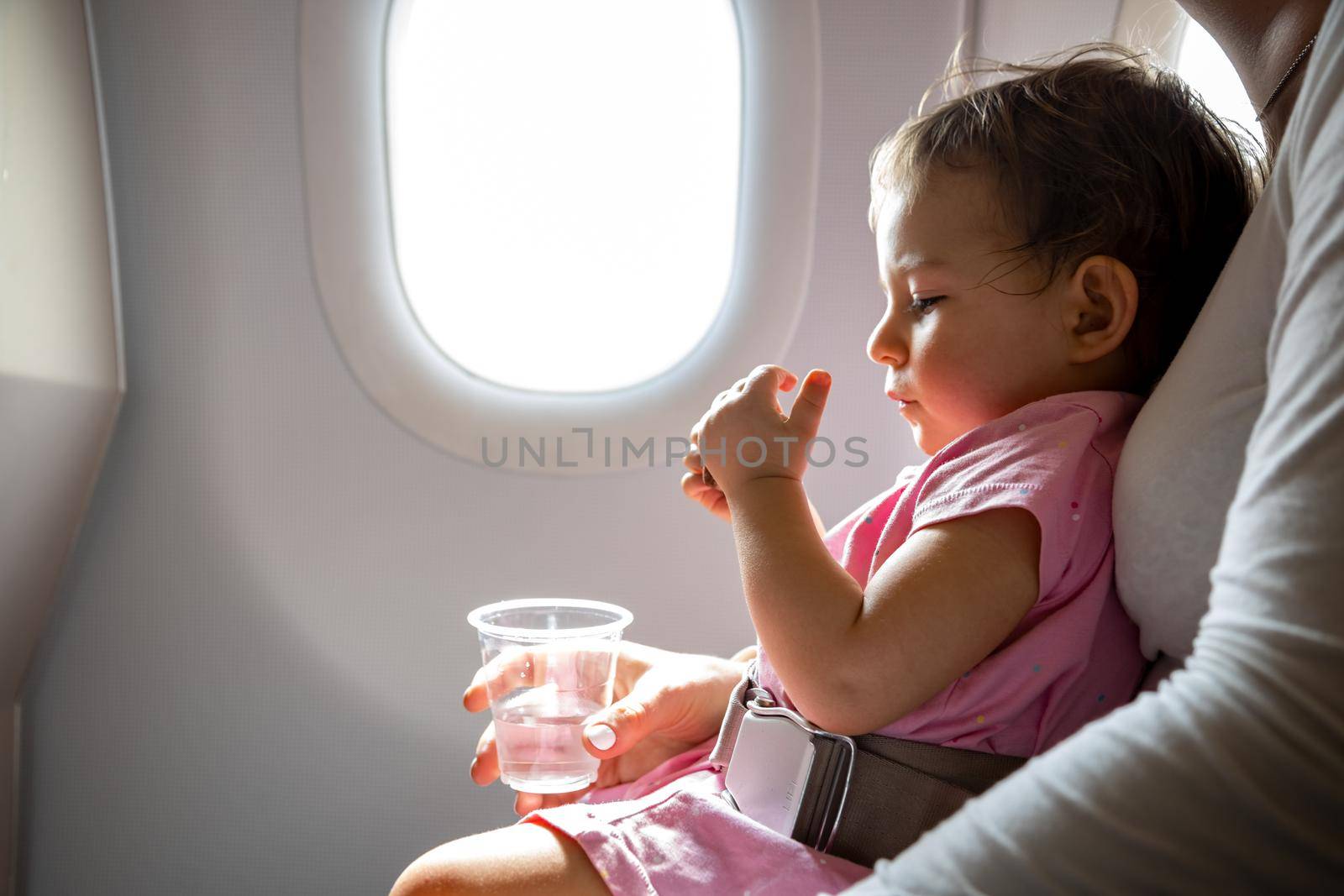 flight with infant. toddler sits on mom’s lap fastened with a special belt in airplane before porthole. by Mariaprovector