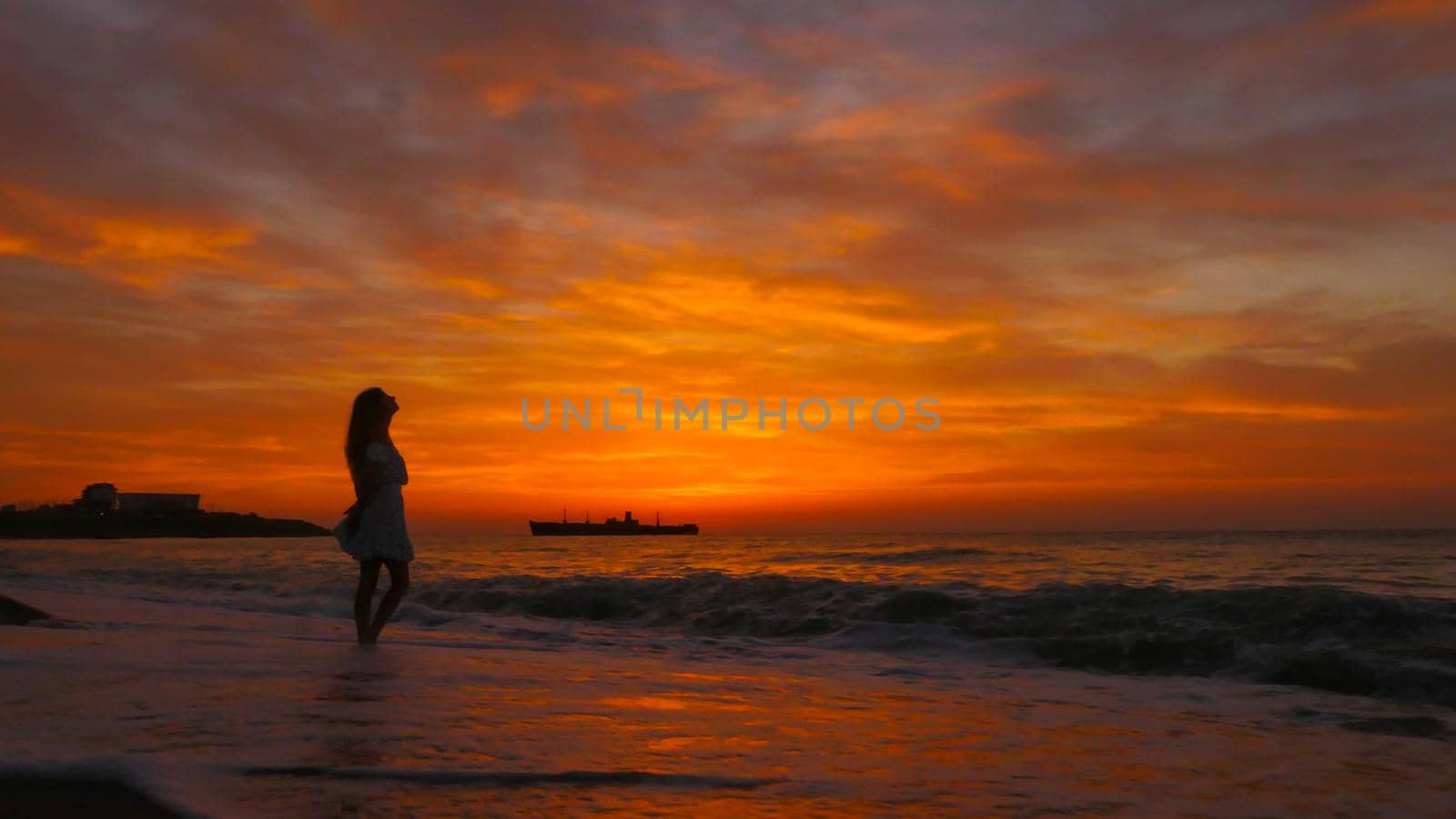 Woman with long hair and summer dress looking at the sea at sunrise. Enjoy the sea breeze in the morning. Colorful summer landscape. A big ship on bacround