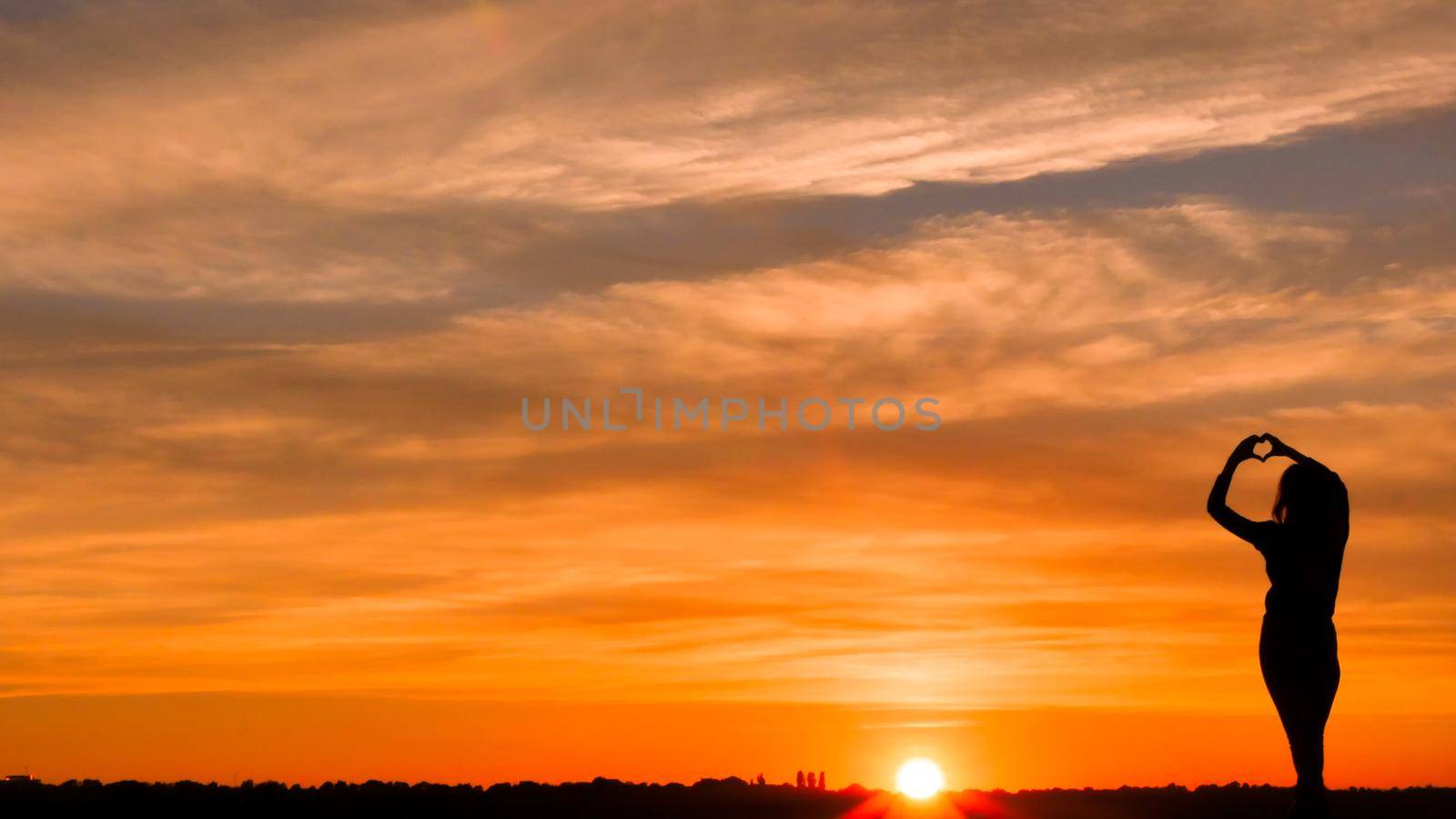 Symbol of love and beautiful things. Woman in love making a heart symbol from fingers in a fire sky background. Silhouette of a happy woman.