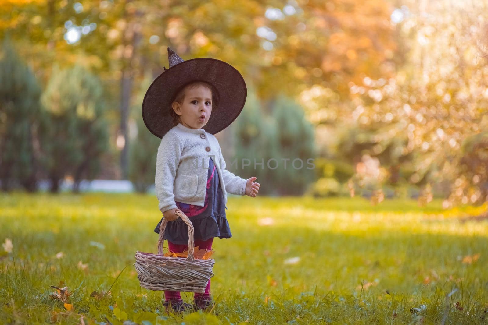 Little girl in witch costume at Halloween in autumn park with basket full of yellow leaves. Childhood, carnival.