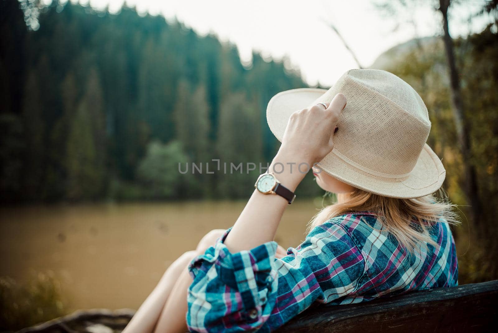 Rear view of a young woman in a denim shirt and hat, sitting on a bench, admires the lake landscape, the pine forest and the relief of the mountains in the background