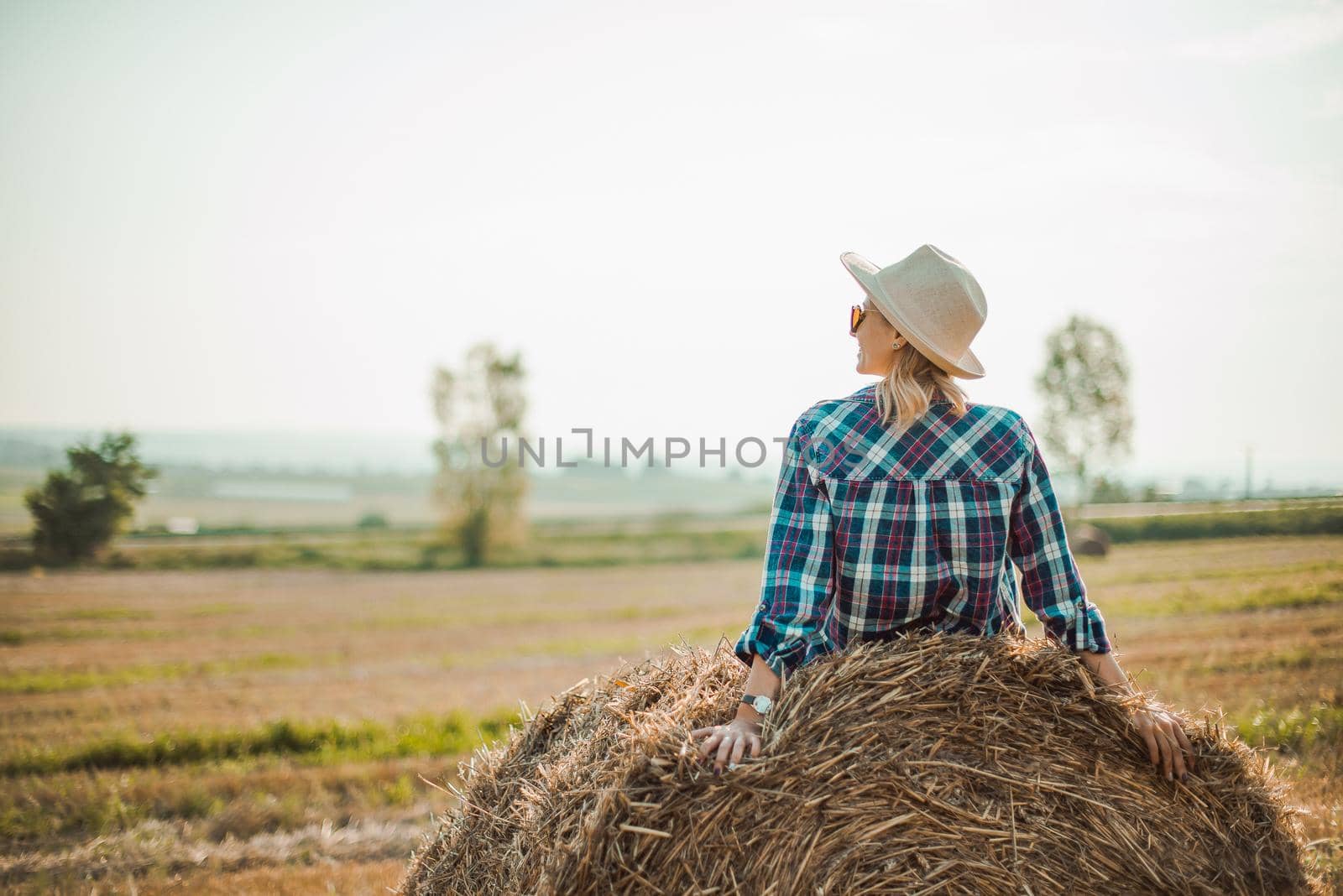 Woman in hat sitting on round bales in the field, enjoying good weather. by RecCameraStock