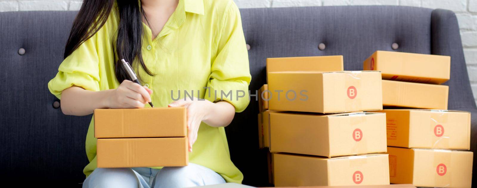 Young asian woman writing address on parcel box at home for delivery order to customer, shipping and logistic, merchant online and seller, business owner or SME, online shopping and e-commerce.