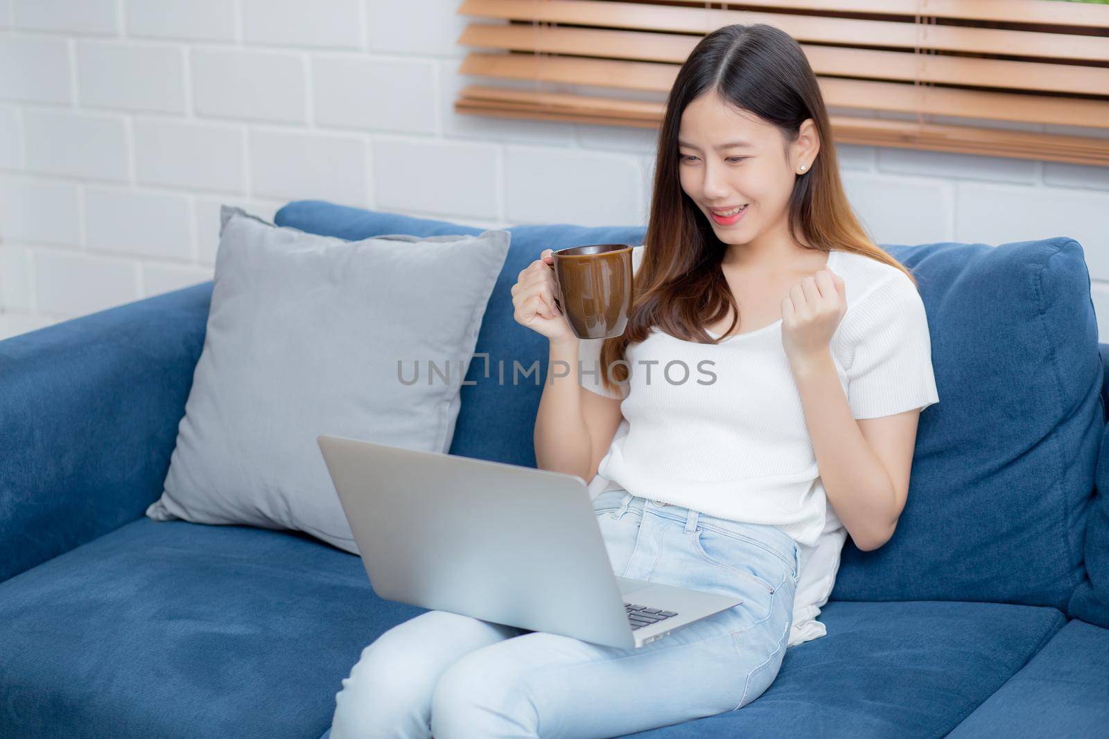 Young asian business woman smile and work from home with laptop computer online on sofa in living room, freelance girl using notebook and drink coffee on couch, new normal, lifestyle concept. by nnudoo