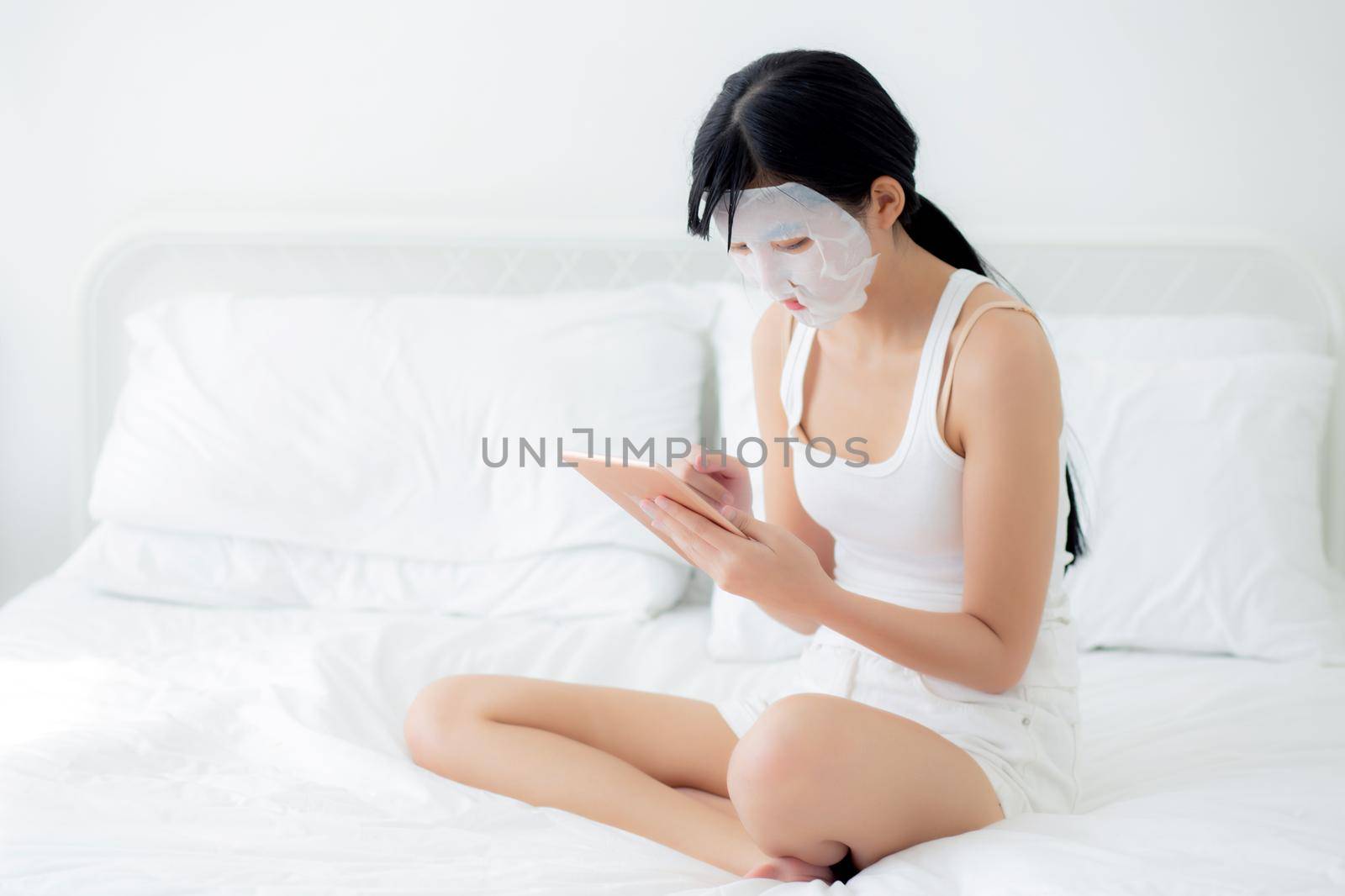 Beautiful young asian woman with sheet facial mask sitting watching tablet computer on bed at bedroom, beauty girl applying face mask for skincare of wrinkle at home, skin care, health and wellness. by nnudoo