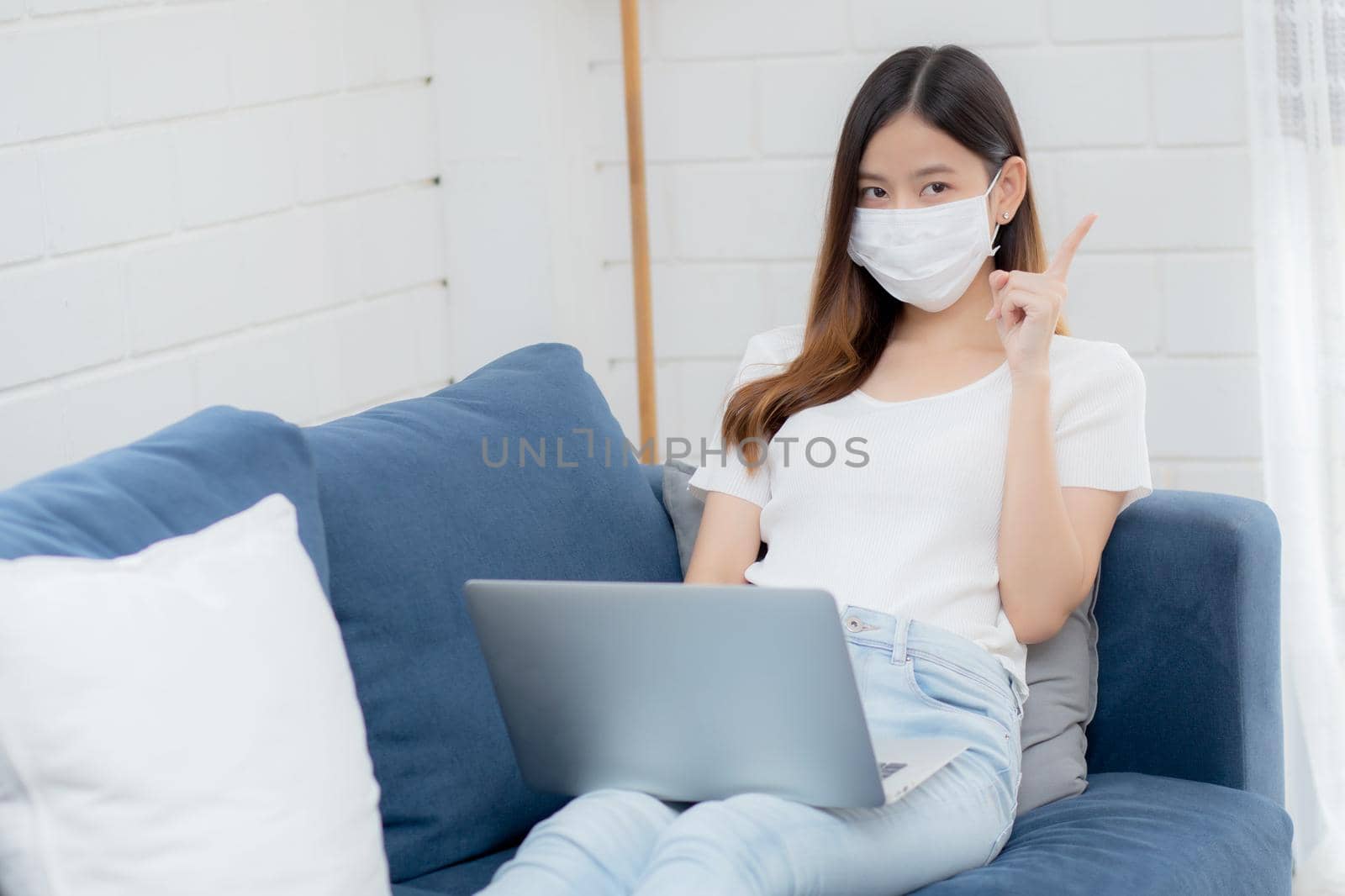 Young asian business woman in face mask think idea and work from home on laptop computer online to internet in living room, girl in medical mask and quarantine for covid-19, stay home, new normal. by nnudoo