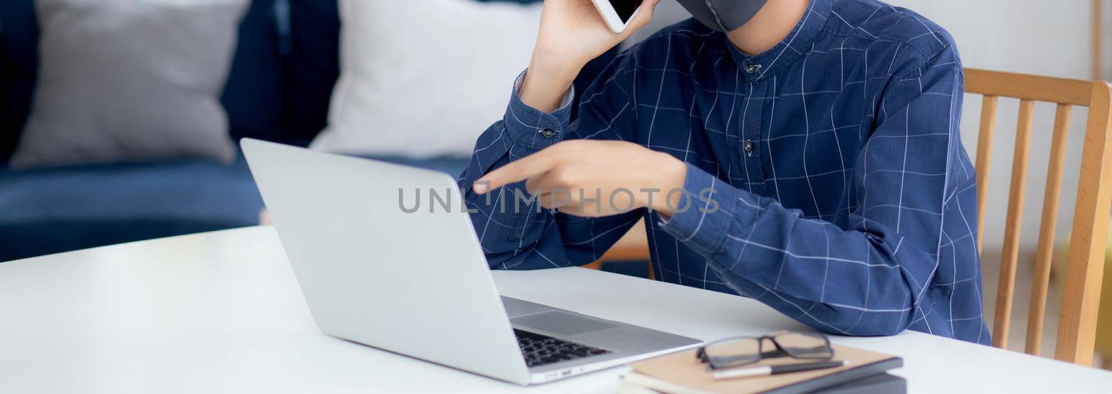 Young asian business man in face mask talking smartphone and work on laptop computer for protect covid-19, businessman quarantine work from home with notebook, social distancing and stay home. by nnudoo