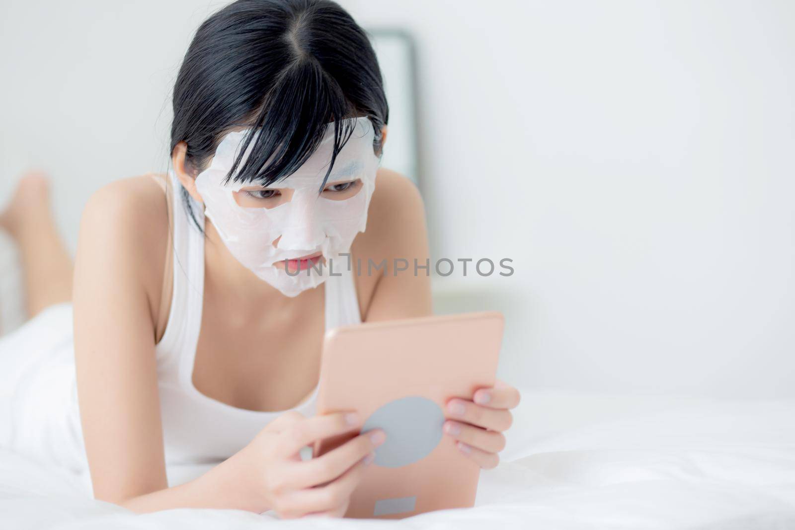 Beautiful young asian woman with sheet facial mask lying watching tablet computer on bed at bedroom, beauty girl applying face mask for skincare of wrinkle at home, skin care, health and wellness. by nnudoo