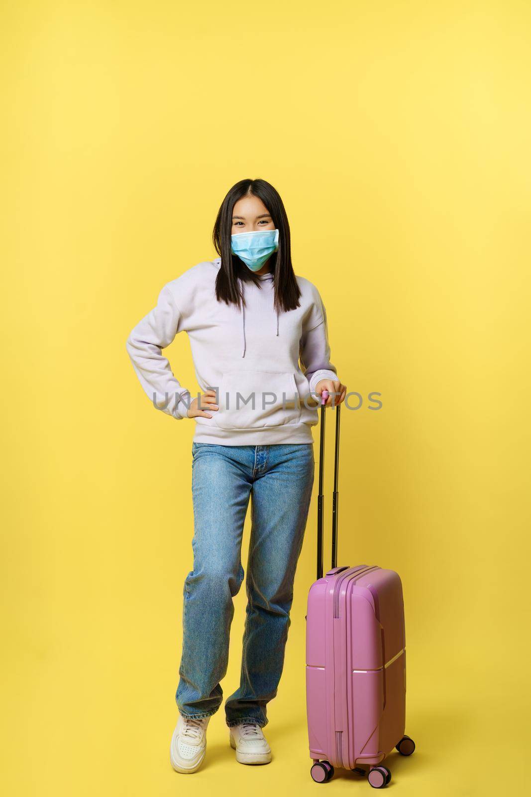 Full length shot smiling korean female tourist in medical face mask, posing with suitcase, going on vacation, standing over yellow background.