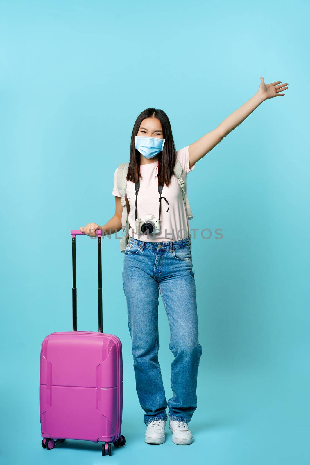 Full length of happy korean woman in face mask, standing with suitcase, raising hand up dreamy, enjoying vacation abroad, standing over blue background by Benzoix