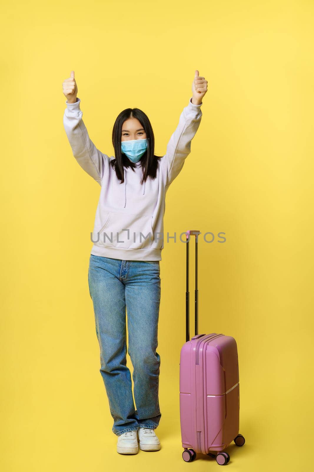 Full length shot of happy asian tourist, girl on vacation in medical face mask, showing thumbs up and standing near suitcase, yellow background.