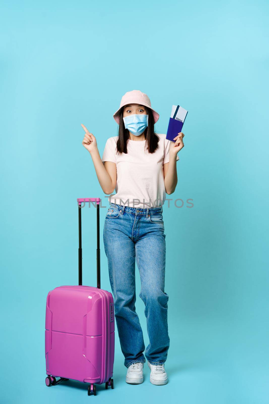 Vertical shot of happy korean woman in medical mask, holding flight tickets and passport, standing with suitcase, dreaming of future vacation, blue background by Benzoix