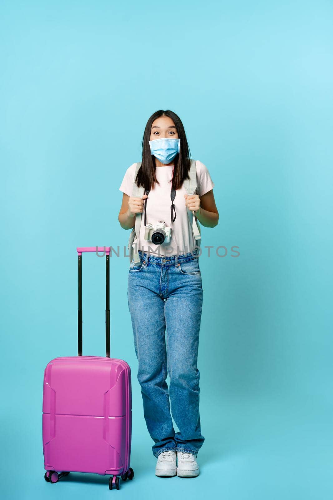 Enthusiastic girl traveller, asian tourist with backpack and suitcase, wearing medical face mask, excited about journey, vacation trip, standing over blue background.