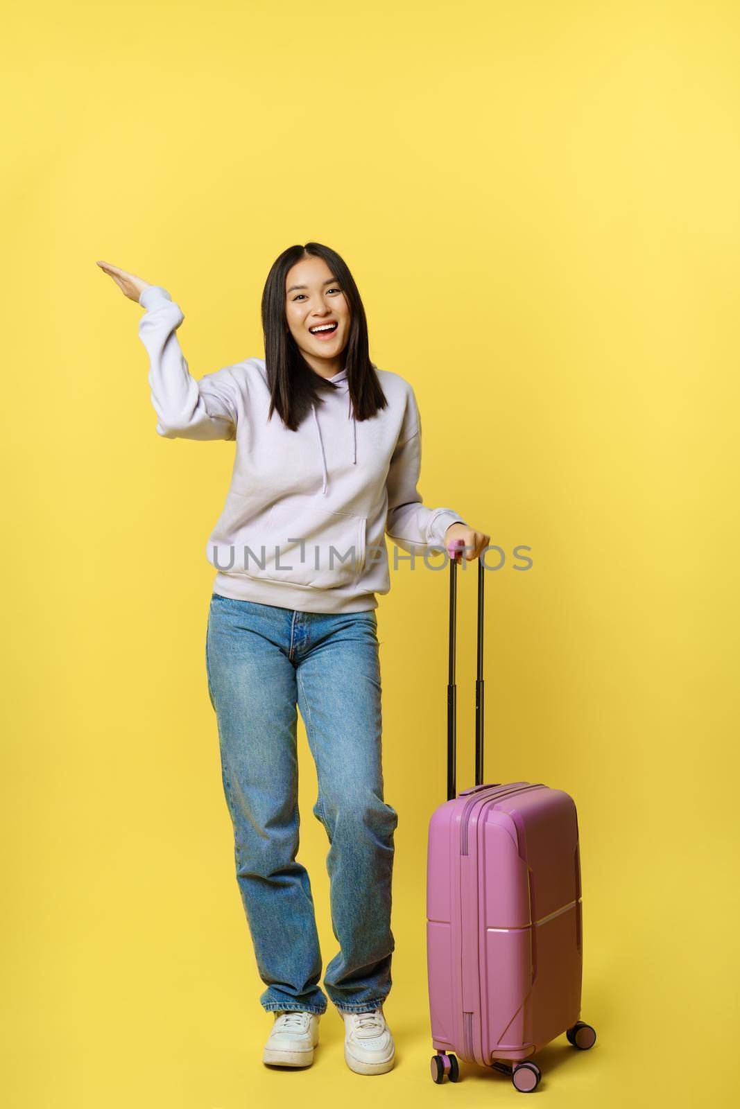 Full length shot happy asian girl going on vacation, tourist with suitcase smiling and looking upbeat, standing over yellow background by Benzoix