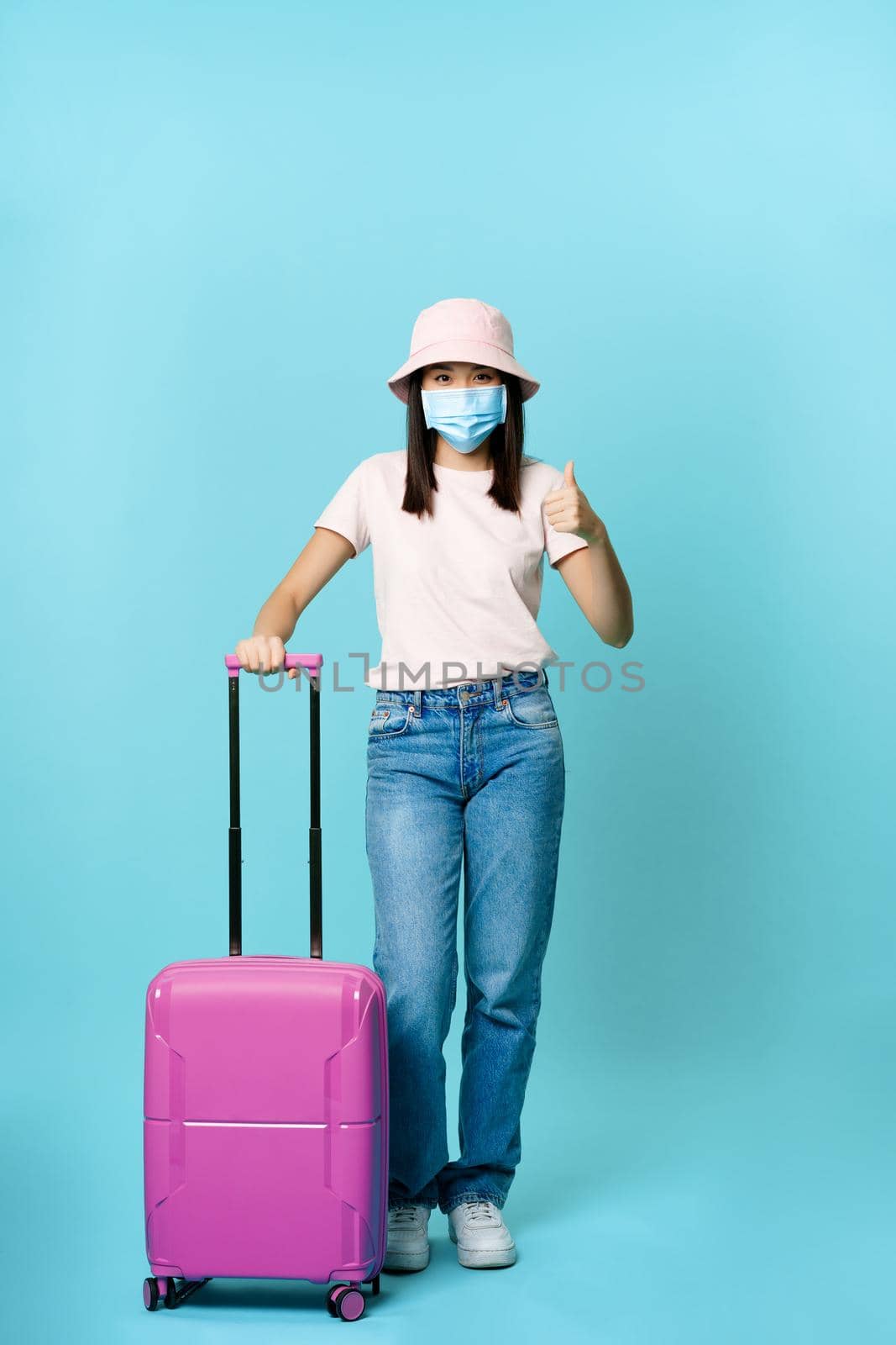 Vertical shot of vaccinated asian girl in medical face mask, going on vacation, standing with suitcase and showing thumb up, blue background by Benzoix