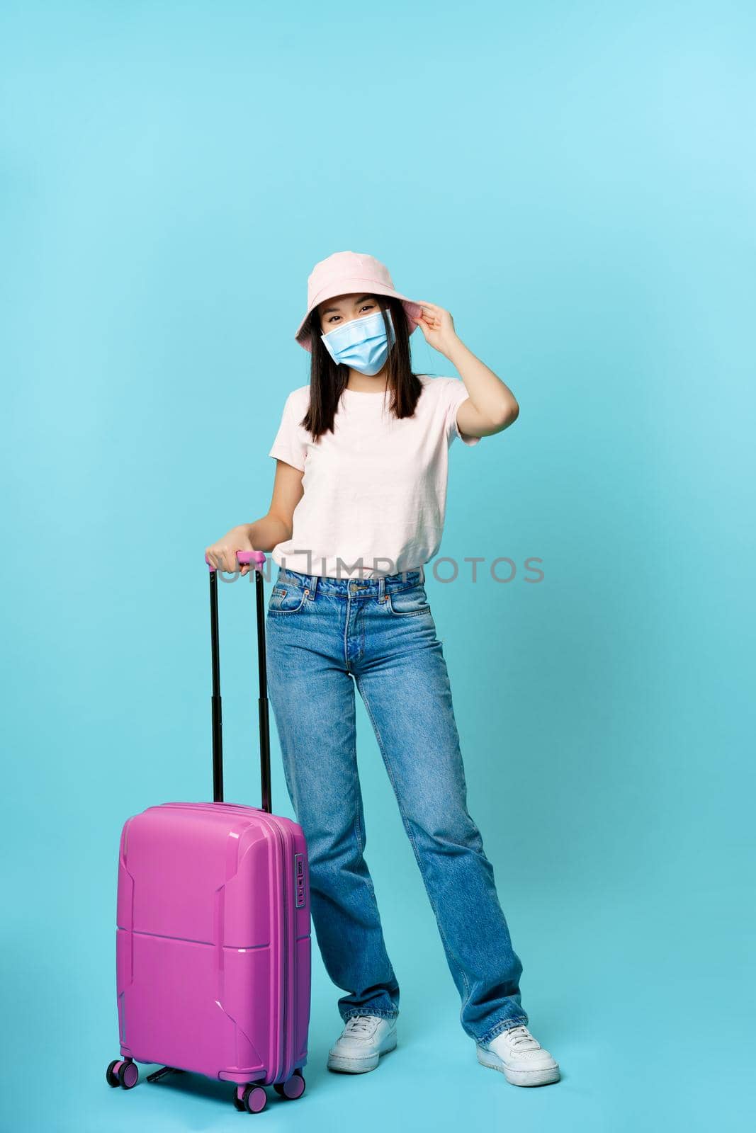 Full length portrait of happy asian woman, traveller with suitcase wearing medical face mask during covid pandemic, going on vacation, blue background.