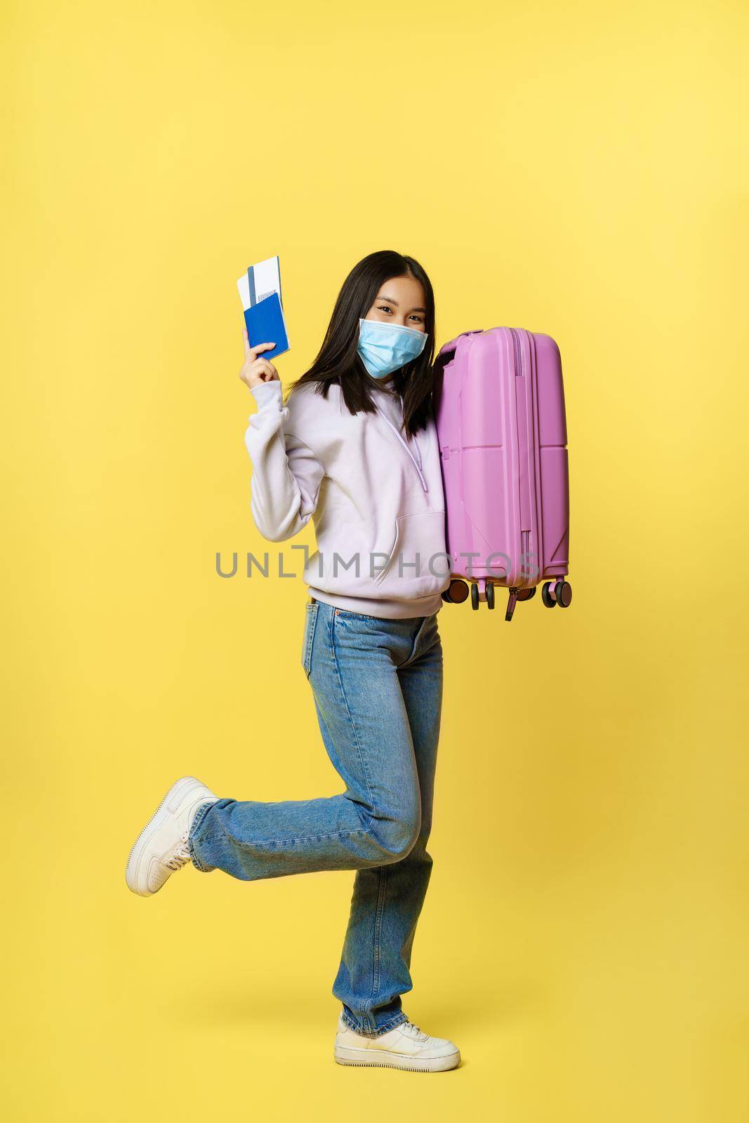 Full length shot cheerful asian female tourist, girl in medical face mask dancing with suitcase and passport with tickets, going abroad on vacation during pandemic.