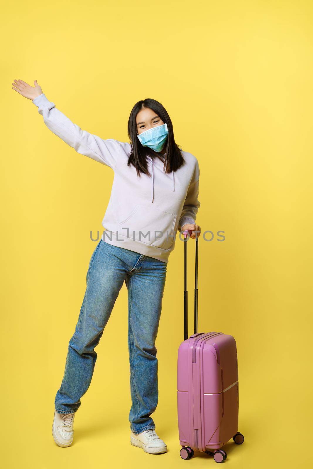 Full length shot of happy korean girl tourist on vacation, posing with suitcase in medical face mask, enjoying travelling, concept of tourism and pandemic healthcare.