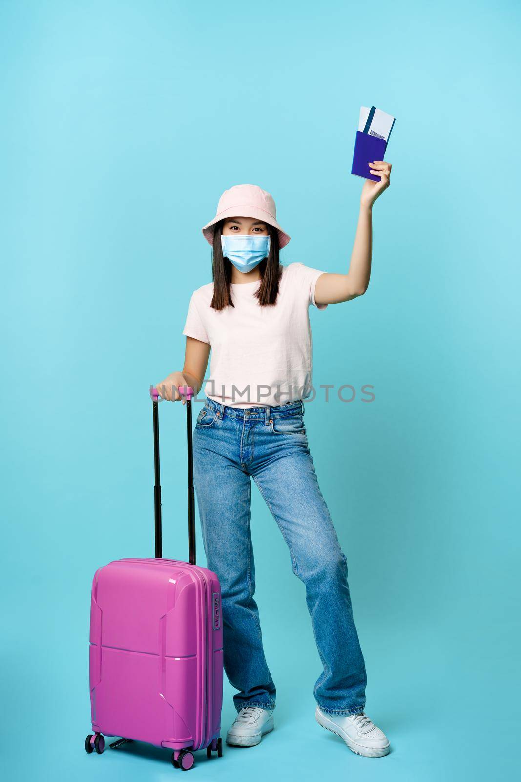 Full length of happy asian tourist, girl travelling abroad, posing with her suitcase in medical face mask, showing passport and tickets, blue background.