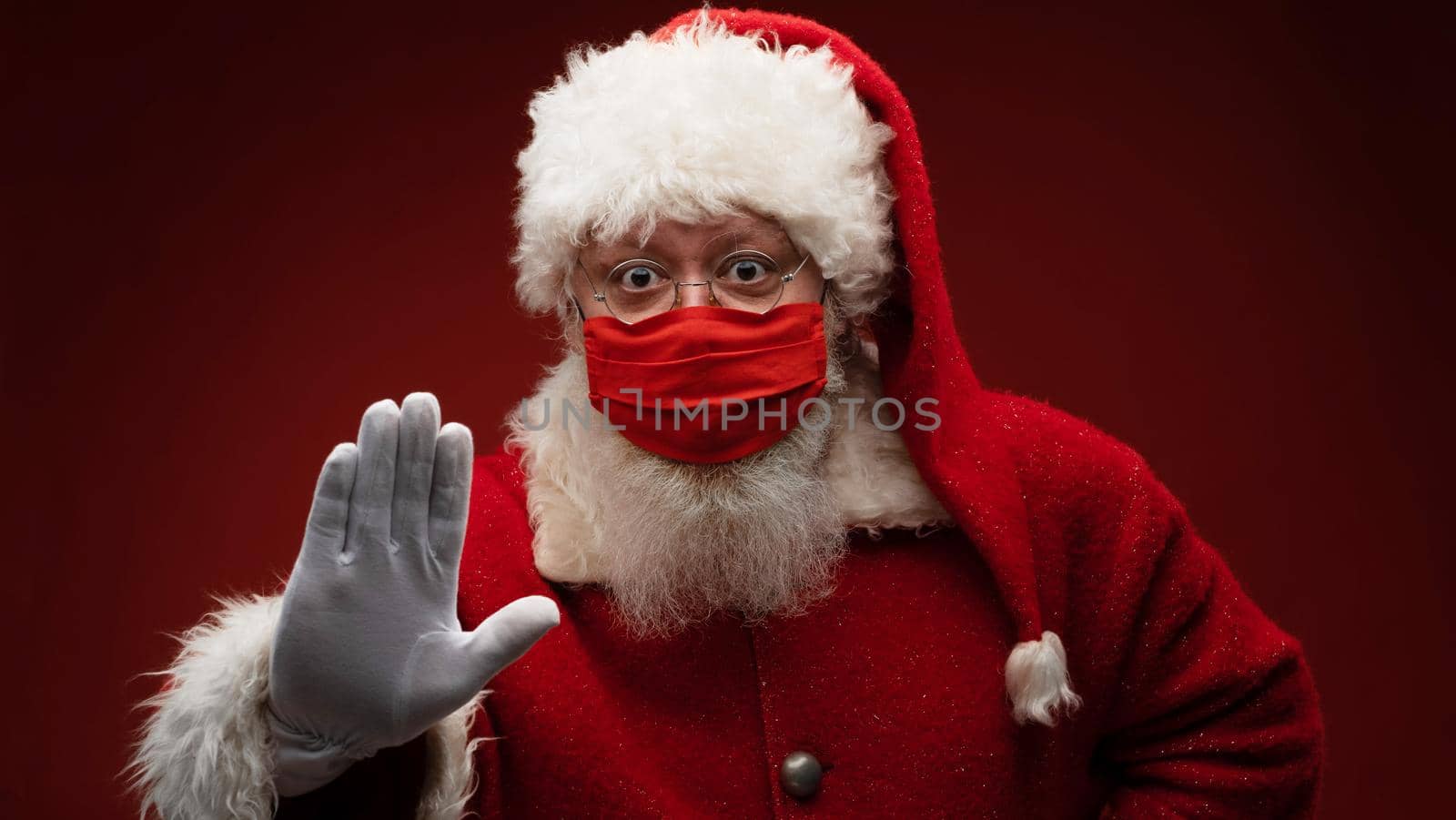 Real Santa Claus on red background, wearing protective mask and with an open hand in front making a pandemic stop sign. Christmas with social distance. Covid-19