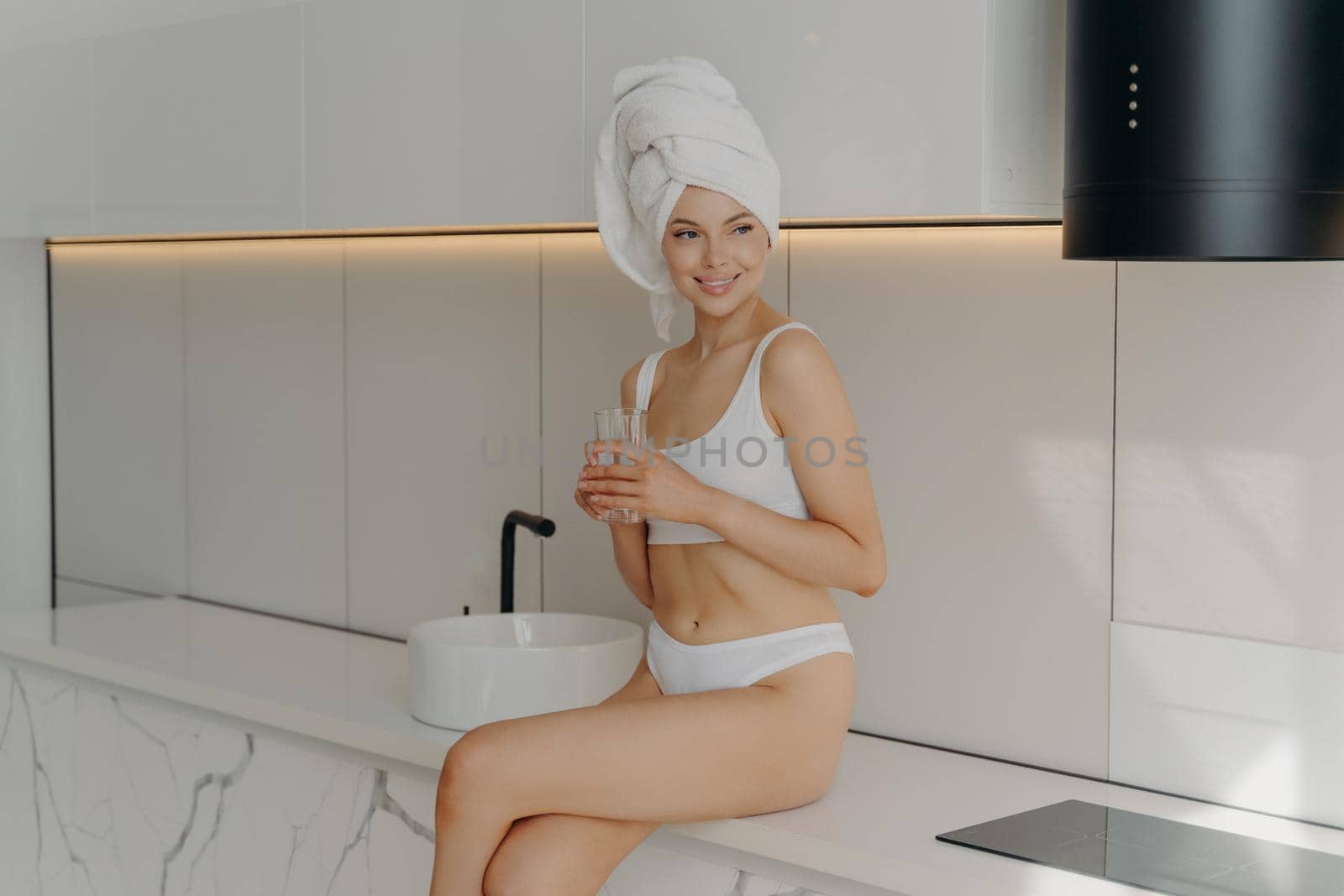 Healthcare concept. Fit slim female sitting on table top in modern stylish light colored kitchen and drinking mineral pure water from glass, looking fresh and clean after morning shower routine