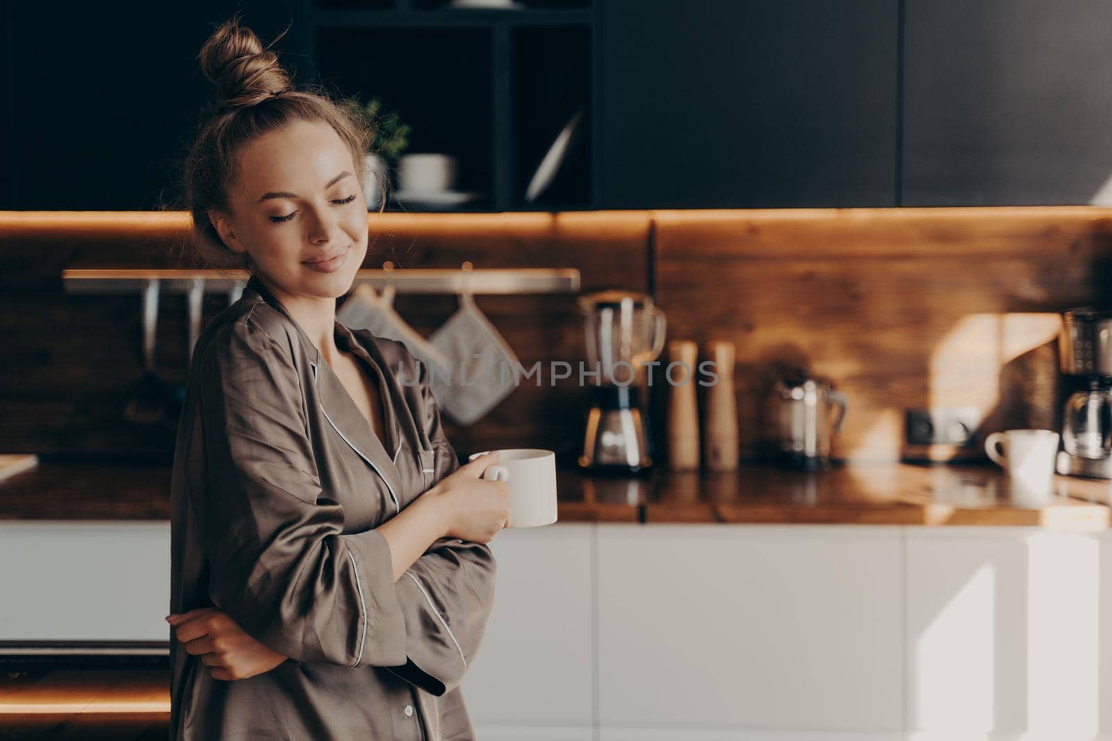 Beautiful young happy woman standing in cozy silk pajama in kitchen with cup of coffee enjoying peaceful early morning with her eyes closed. Leisure time at home concept