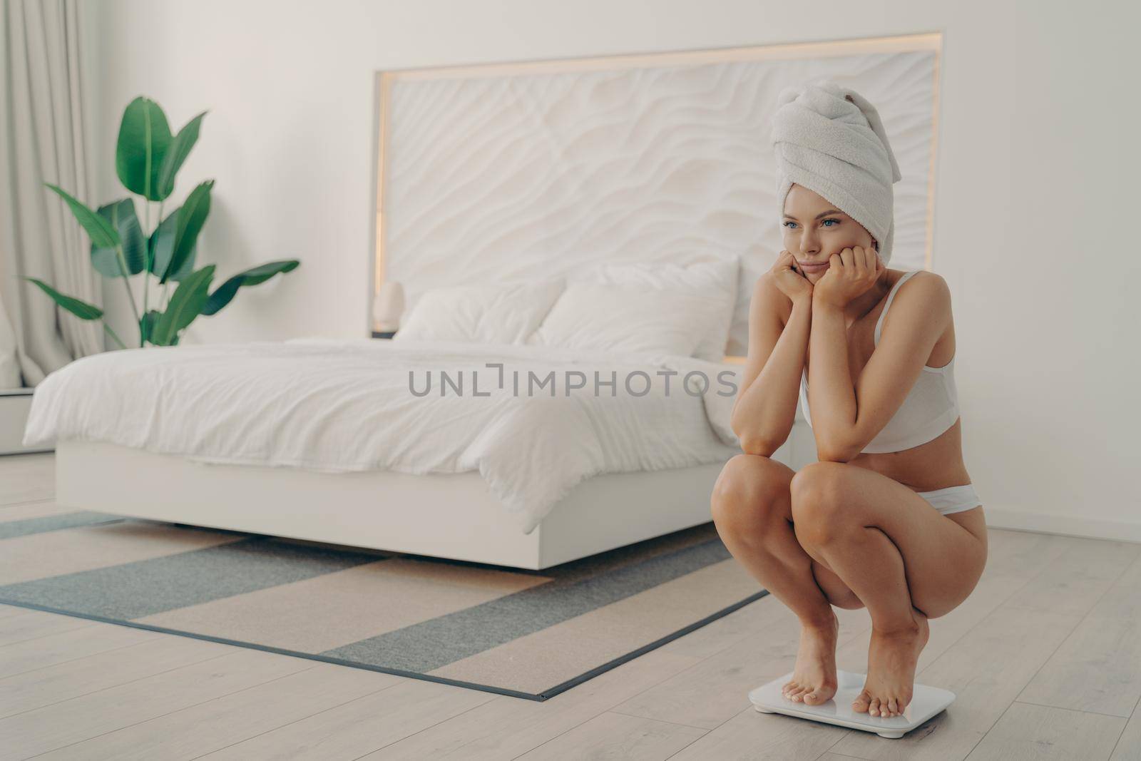 Young sad and unhappy girl in underwear barefoot in underwear squatting on electronic smart scales by vkstock